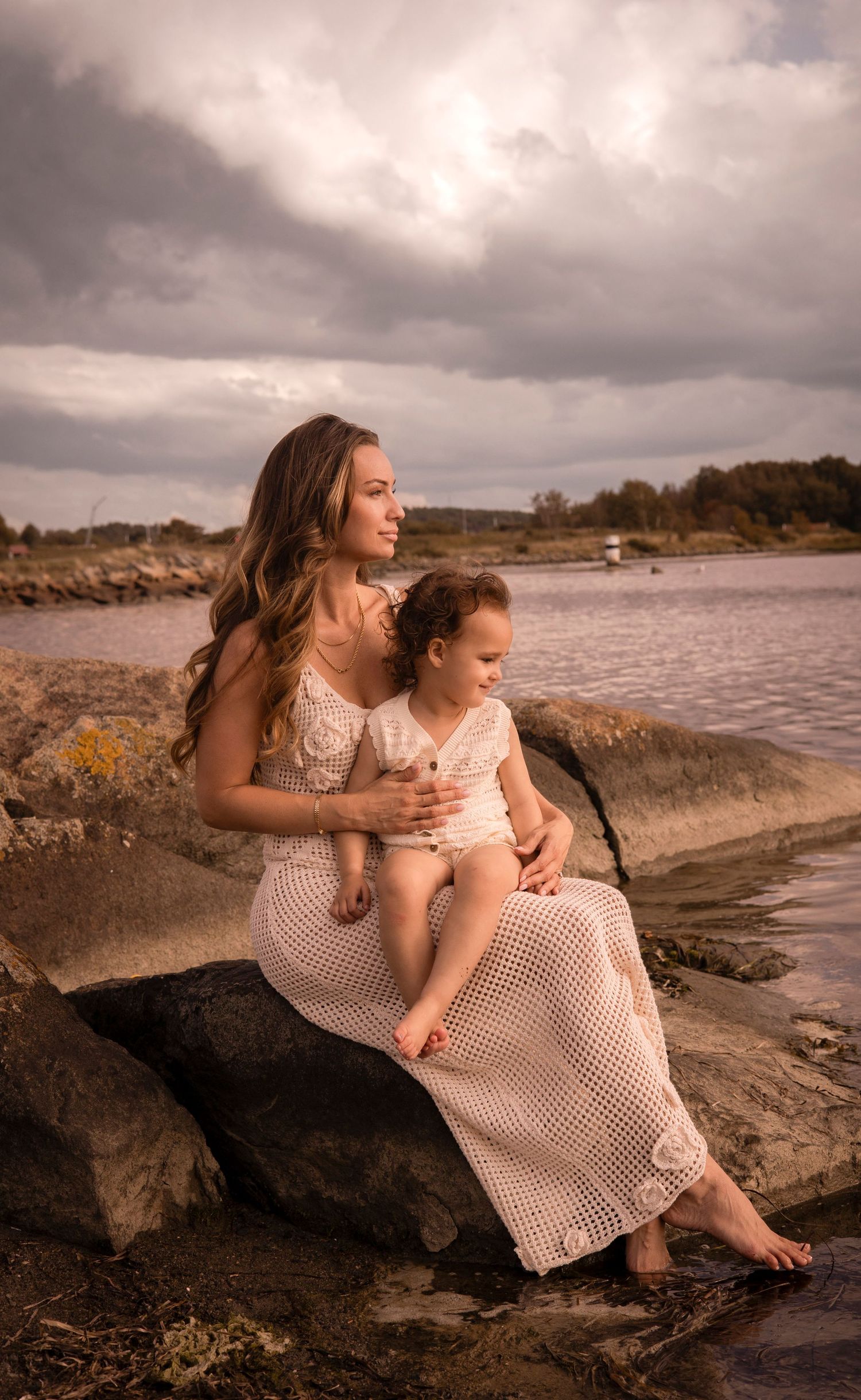 mamma sitter med sitt barn på en sten bland klippor. de kollar ut mot havet. båda är klädda i ljusa färger.