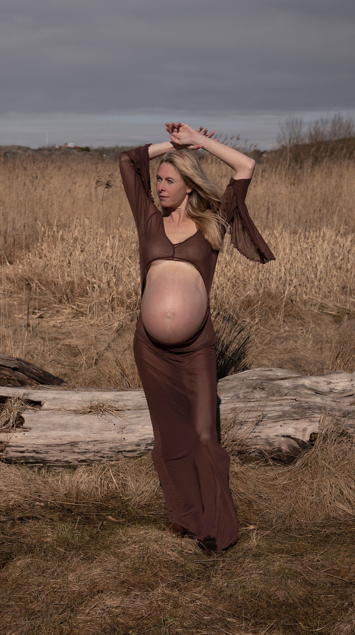 gravid kvinna står på en äng i bakgrunden syns en stock. hon har händerna över huvudet och på sig en brun klänning.