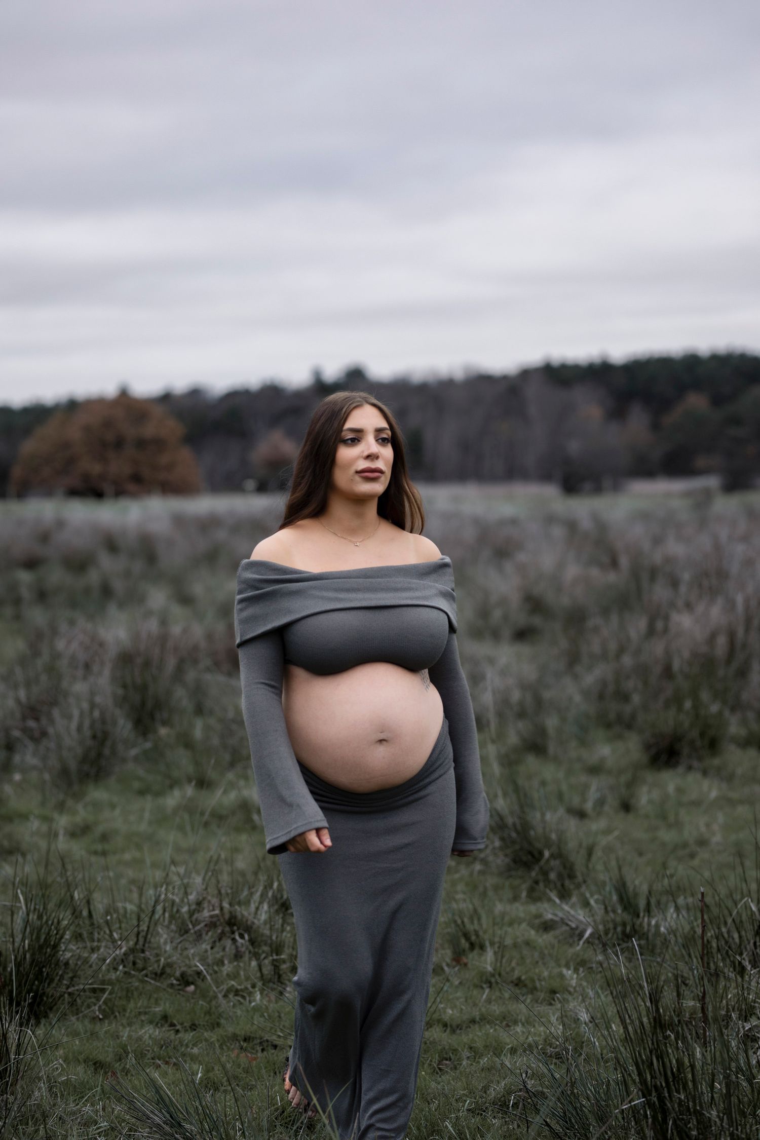 gravid kvinna står på en åker. på sig har hon ett grått stickat set bestående av en långärmad topp och en kjol.