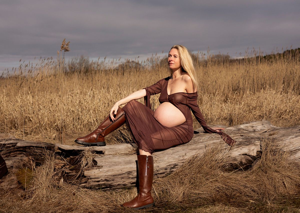gravid kvinna sitter på en liggande stock. på sig har hon en brun klänning och bruna boots. hon har ena benet upp på stocken.