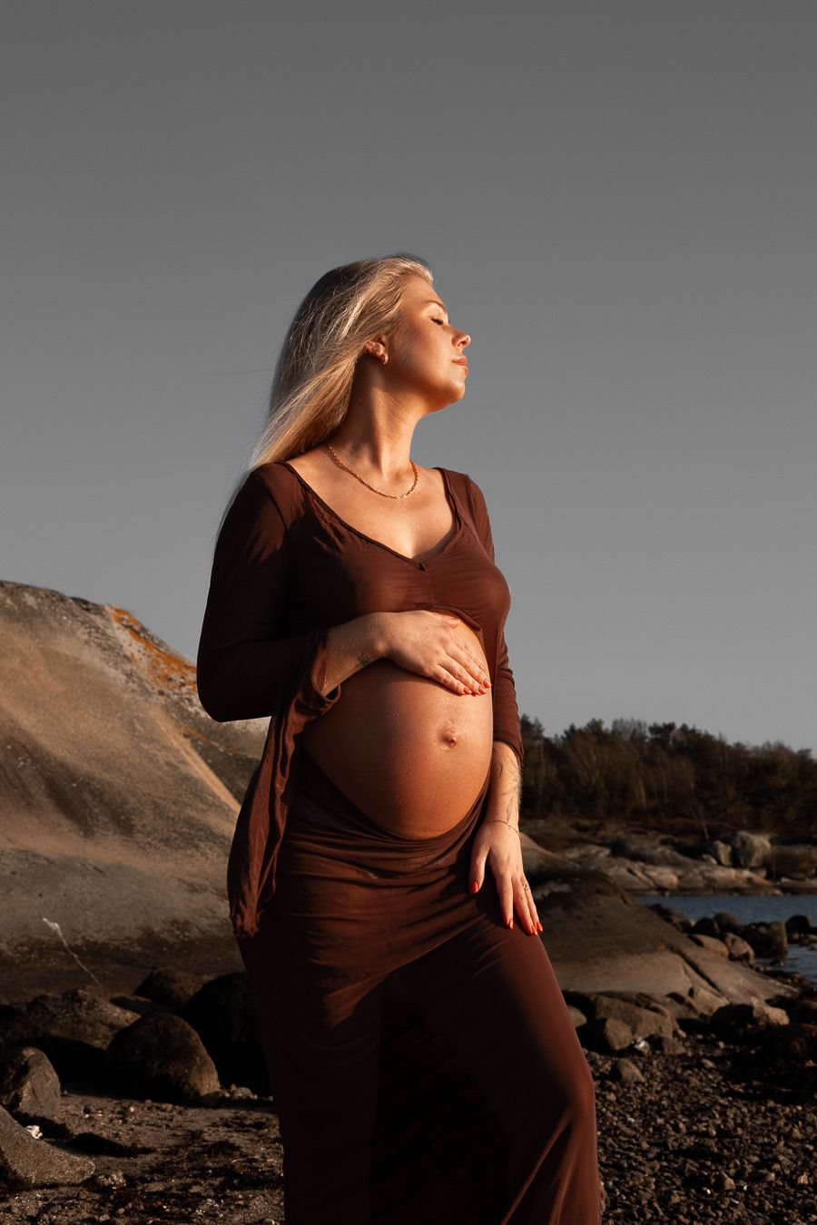 gravid kvinna står på en strand i solens gyllene ljus. hon håller ena handen på sin gravidmage. i bakgrunden syns en klippa.