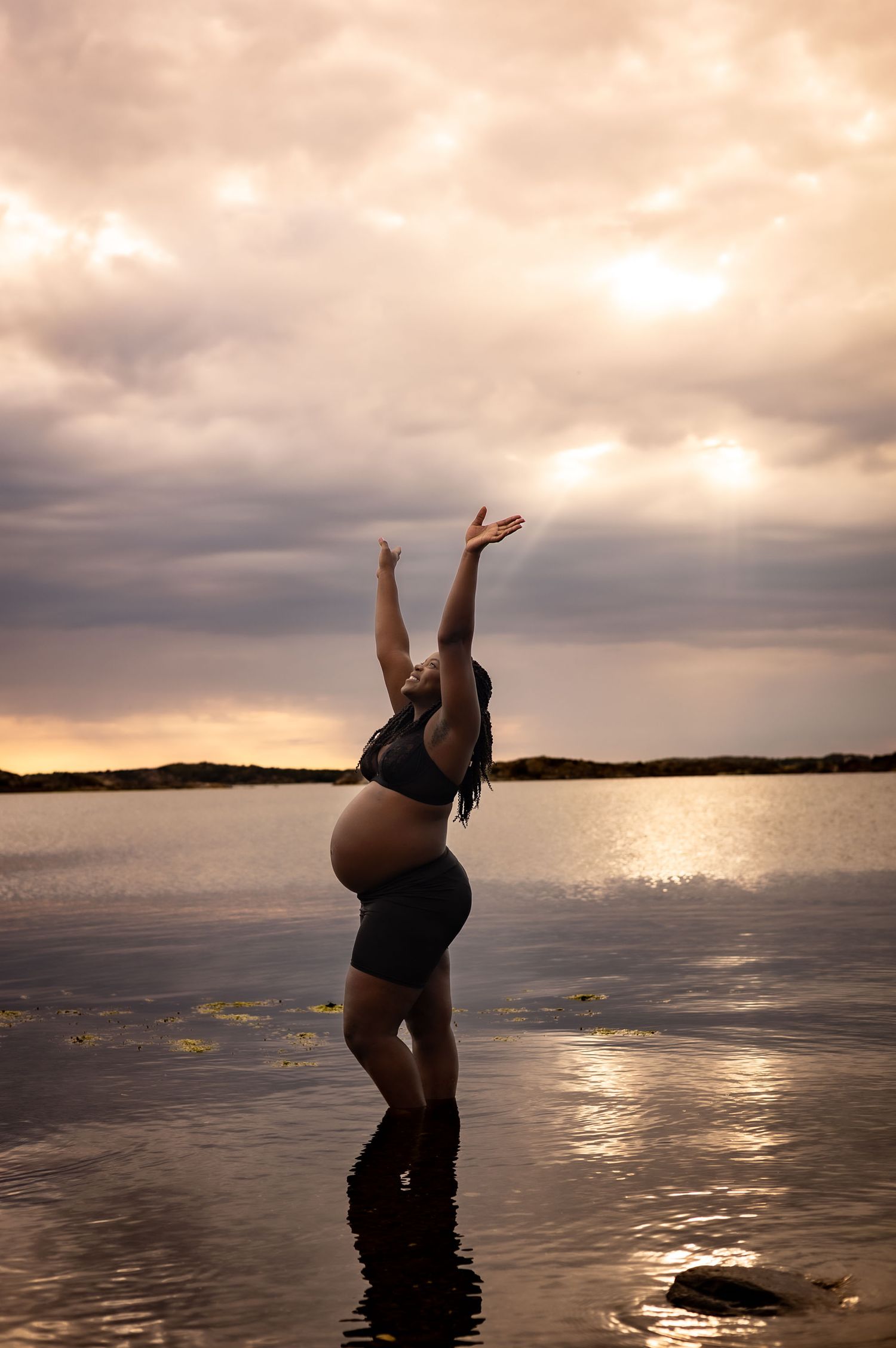 gravid kvinna står i vattnet i bakgrunden går solen ned, hon har ett par svarta shorts och en svart topp.