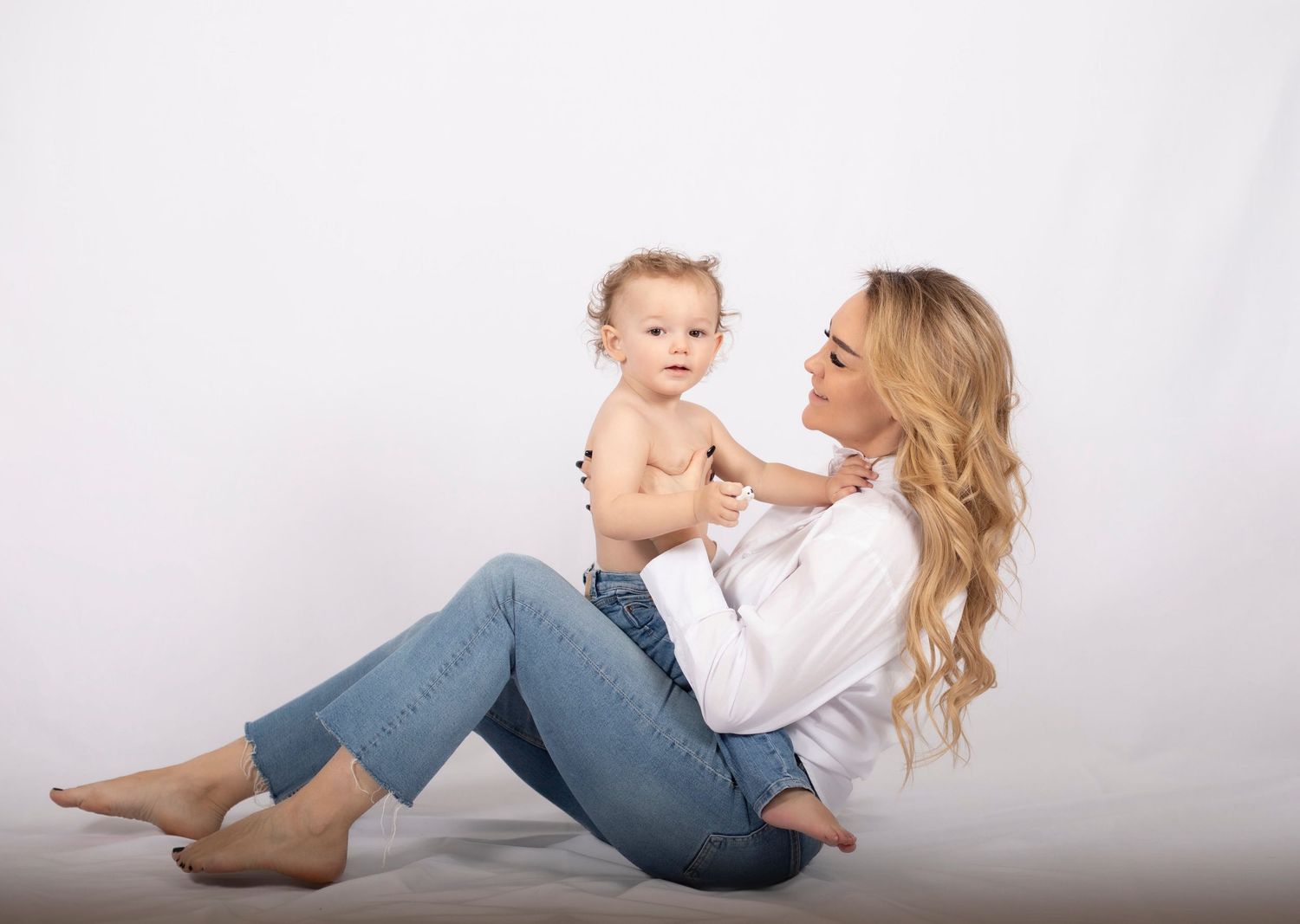 mamma och barn sitter i en vit studio. barnet sitter i mammans knä. på sig har de jeans och mamman har en vit skjorta.