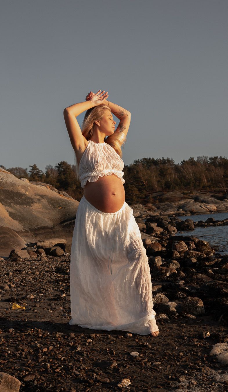 gravid kvinna står på en strand. hon har ett vitt set på sig och håller armarna över huvudet och kollar ut mot havet.