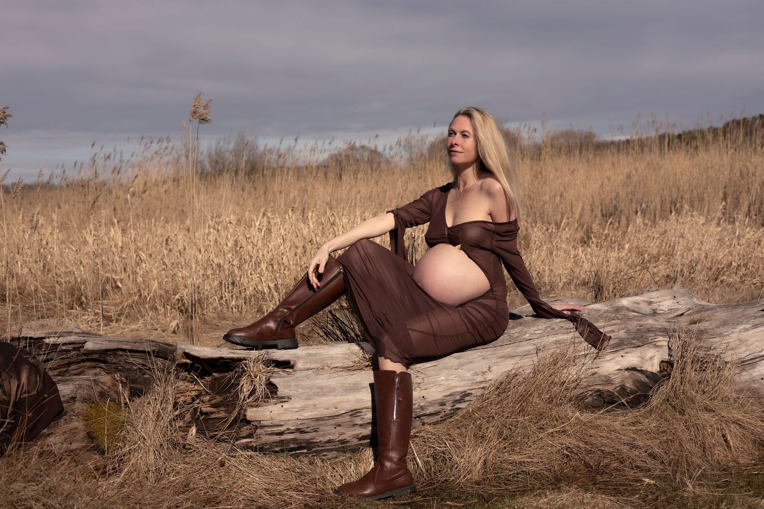 kvinna sitter på en stock, hon är gravid. på sig har hon en brun klänning med hål för magen och bruna boots.