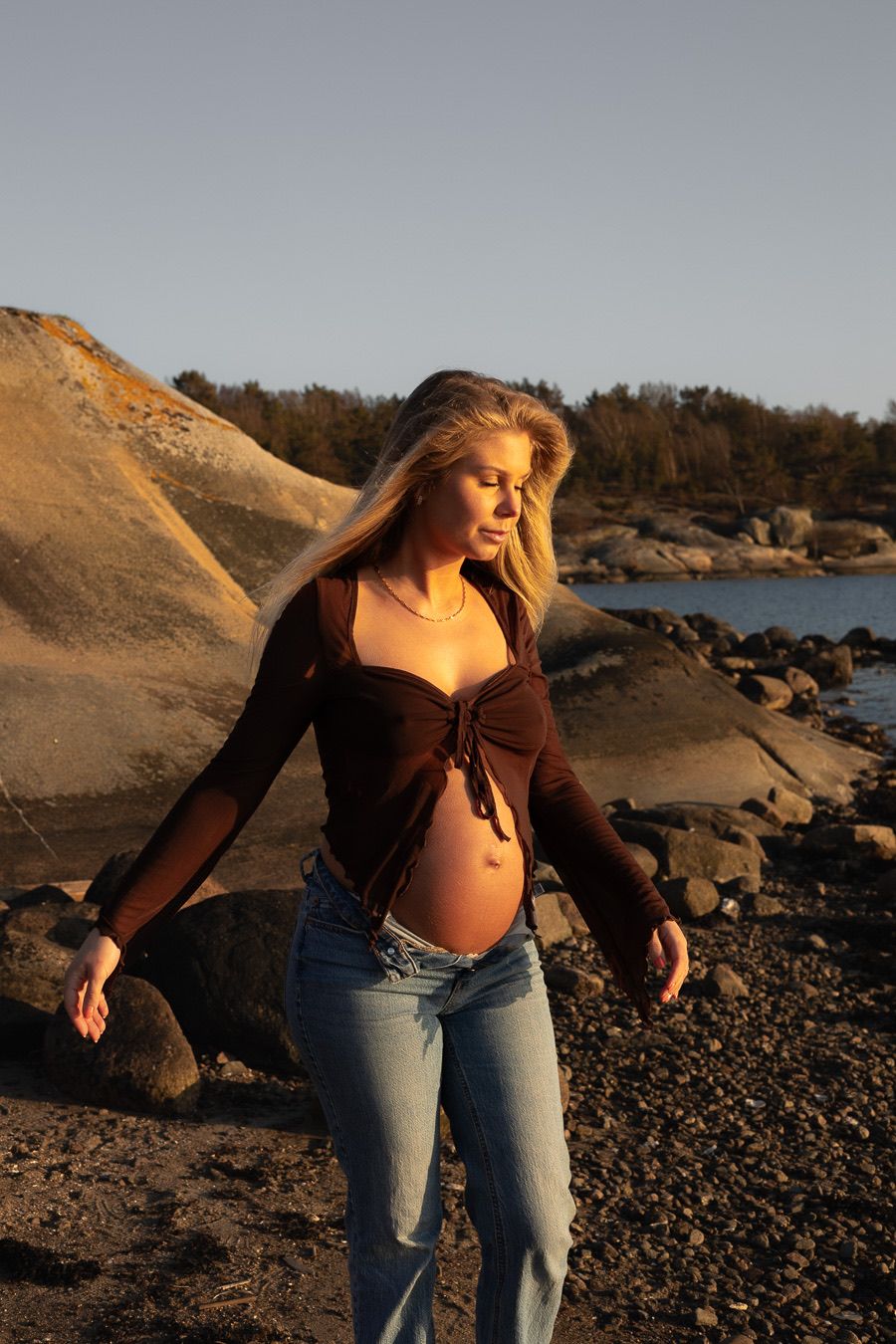 Gravid kvinna står på en strand. på sig har hon en brun topp och ett par jeans.
