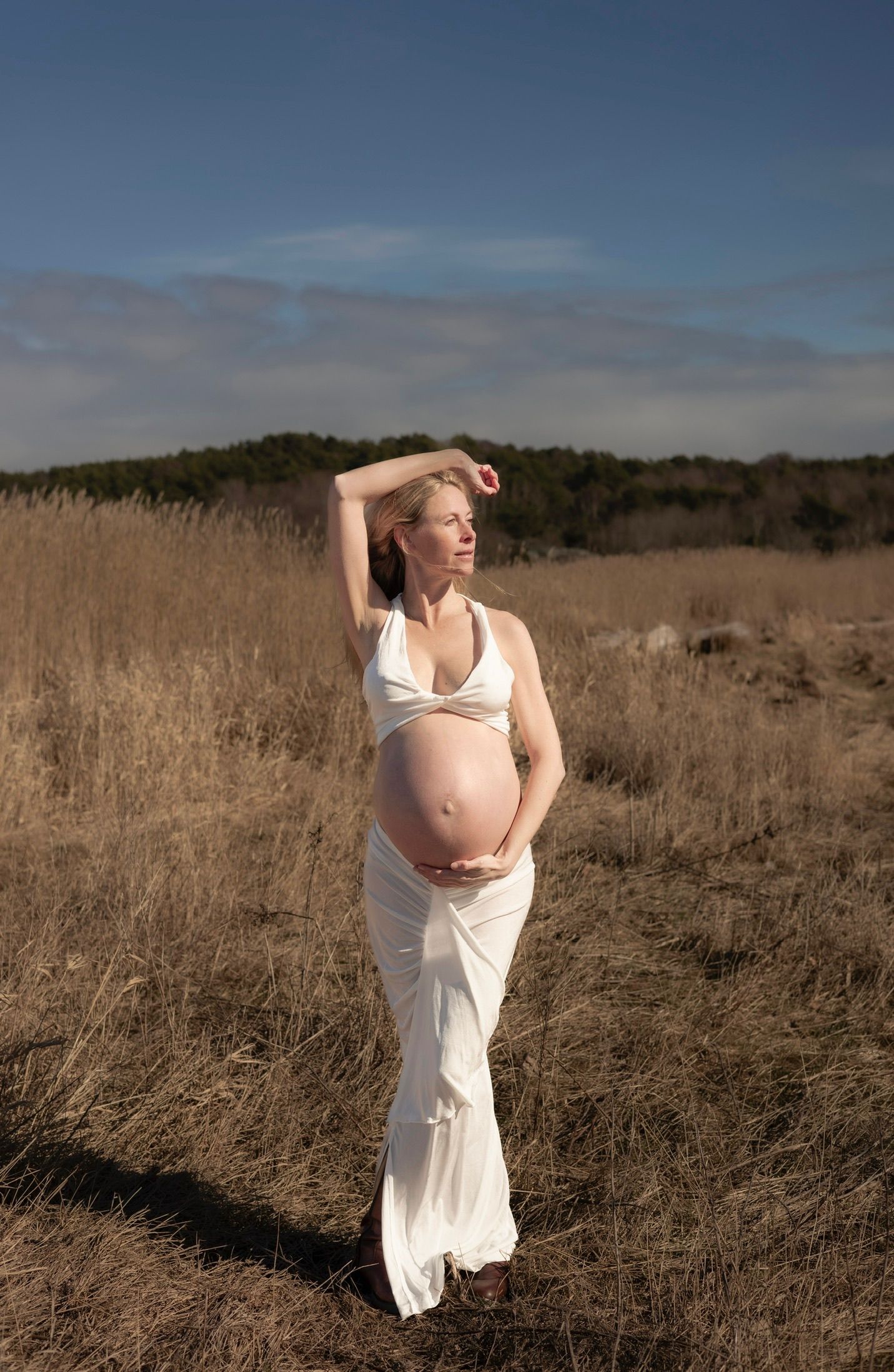 gravid kvinna står på en åker. hon har ena armen ovanför huvudet och andra armen runt magen. hon har ett vitt set på sig.