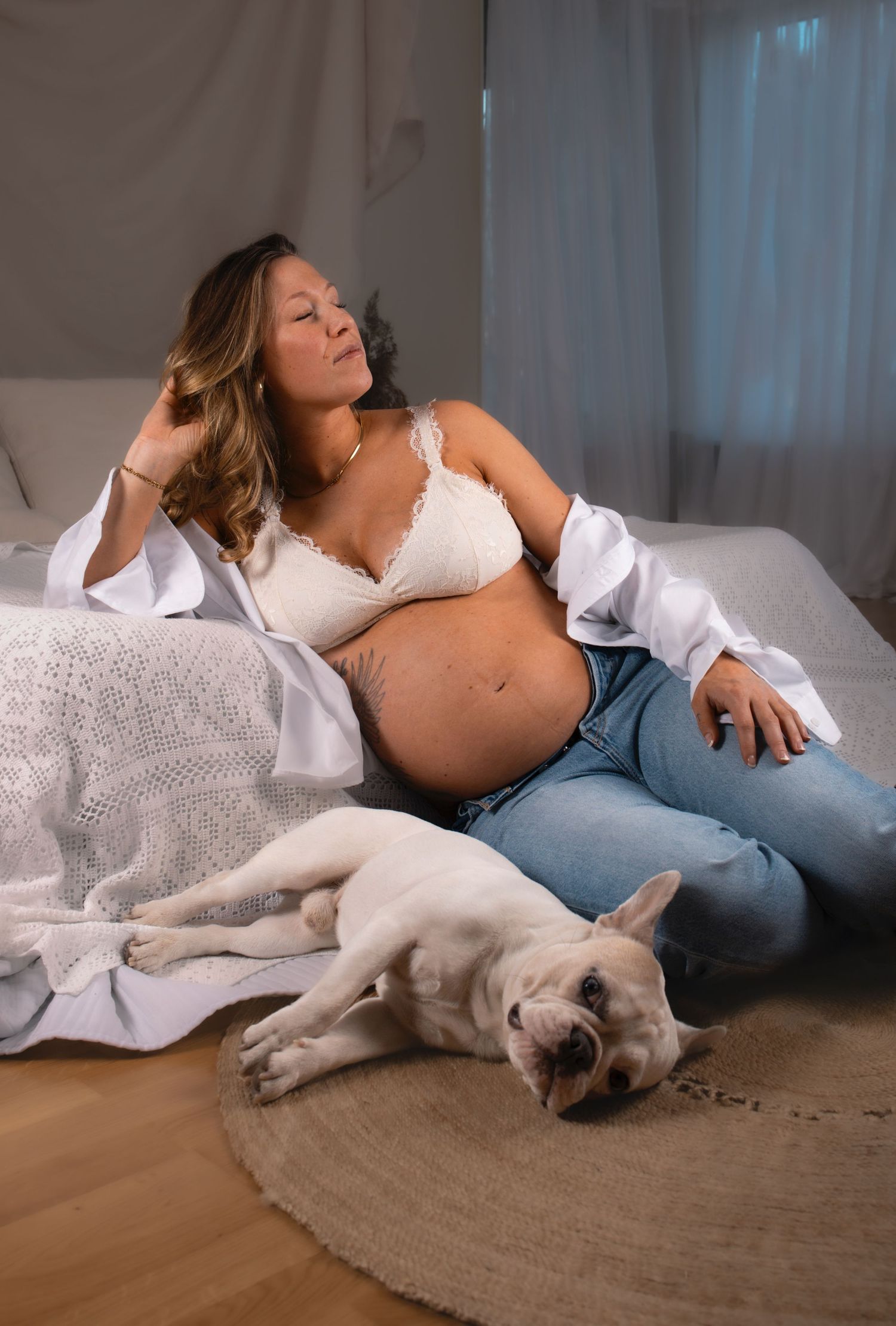 gravid kvinna sitter lutad emot en säng. framför henne ligger en hund. hon har jeans, en vit topp och en vit skjorta på sig.