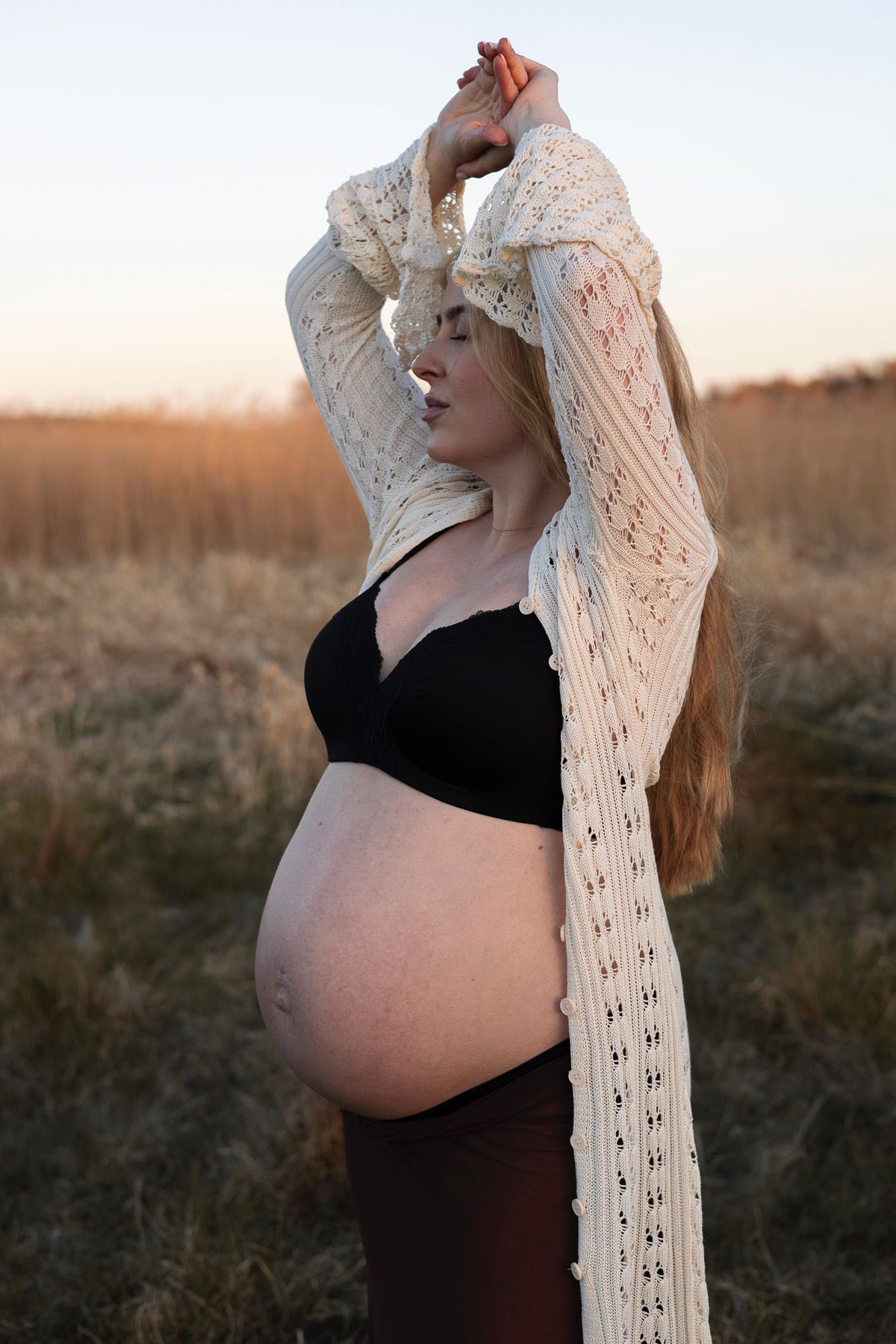 gravid kvinna står med armarna upp. på sig har hon en svart topp, bruna byxor och en vit stickad kofta.