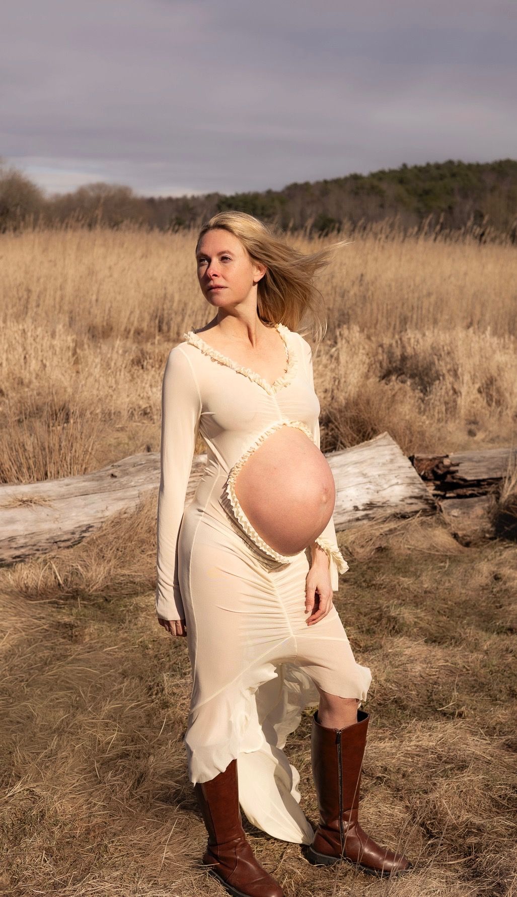 Gravid kvinna står på en äng. på sig har hon en ljus klänning med ett hål för magen och ett par bruna boots. hennes hår blåser i vinden.