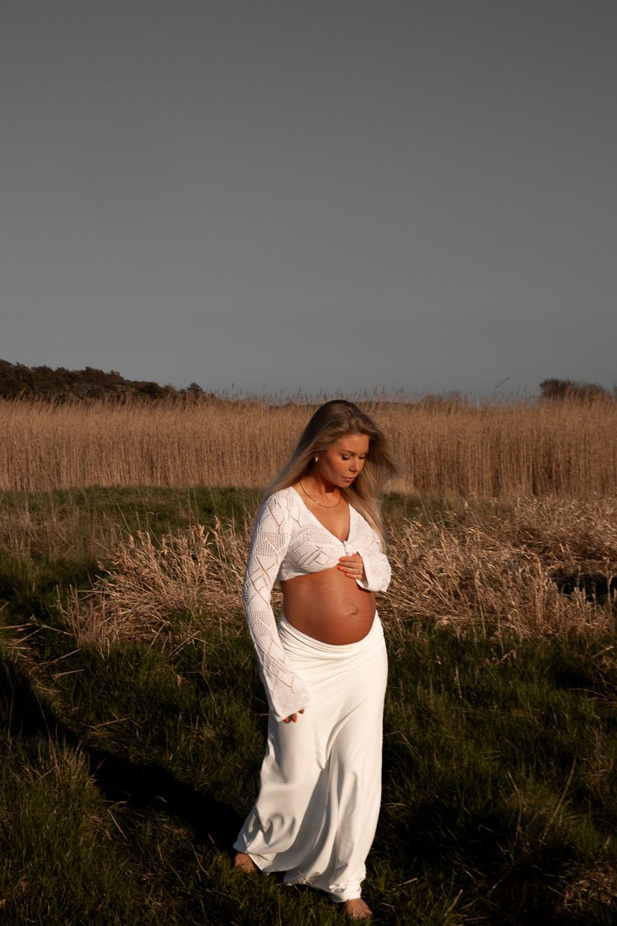 gravid kvinna går på en åker. solen lyser på henne. hon har ett vit set med en långärmad tröja och en vit kjol.