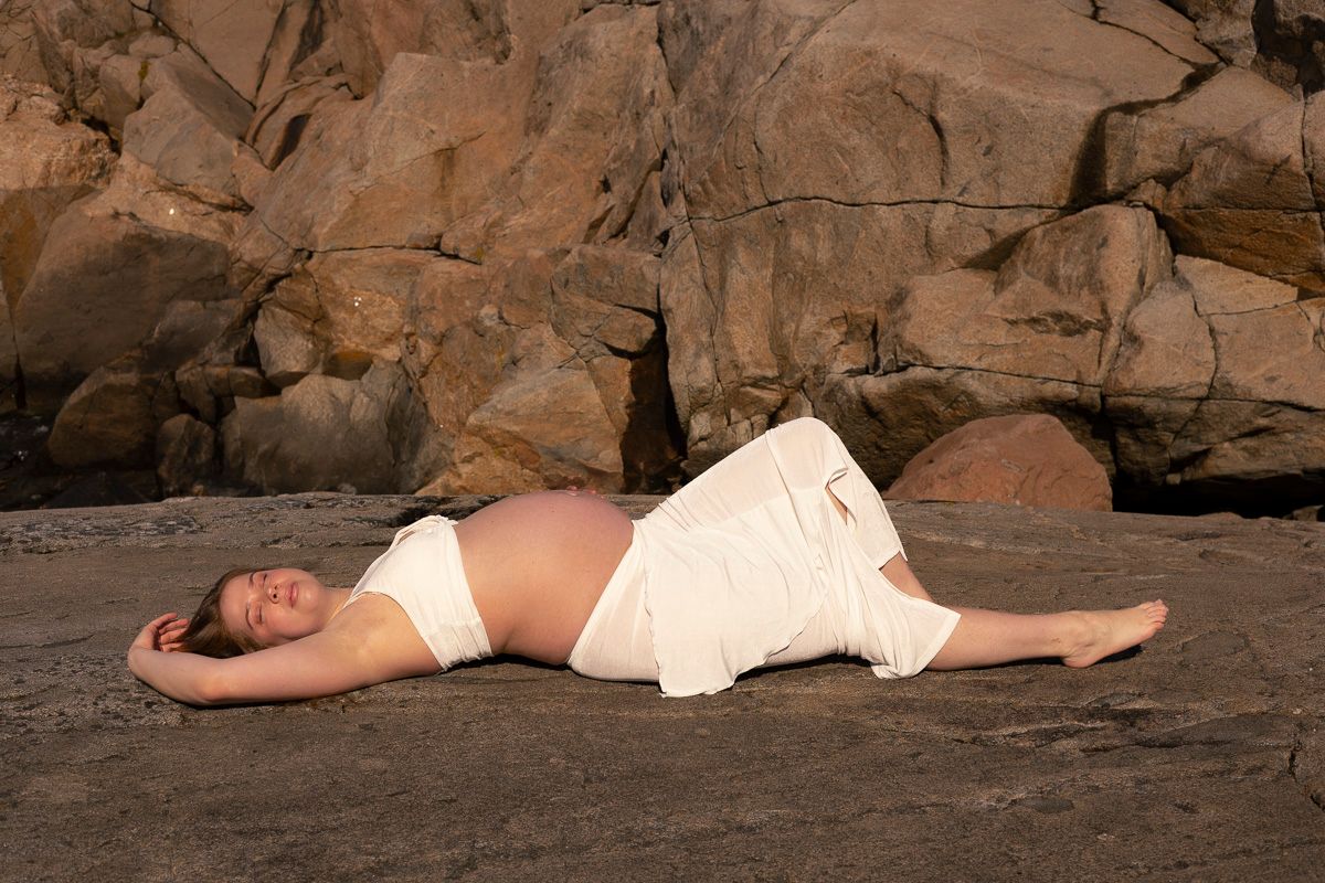 gravid kvinna ligger ner på en klippa. på sig har hon ett vitt set bestående av en kjol och en topp.