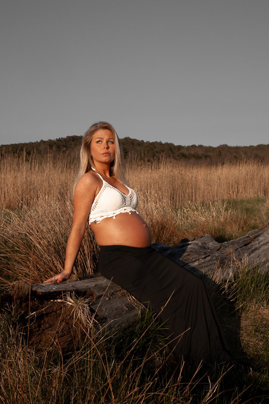Kvinna som är gravid sitter på en stock. hon har armarna bakom sig på stocken lutandes. på sig har hon en vit topp och en svart kjol.