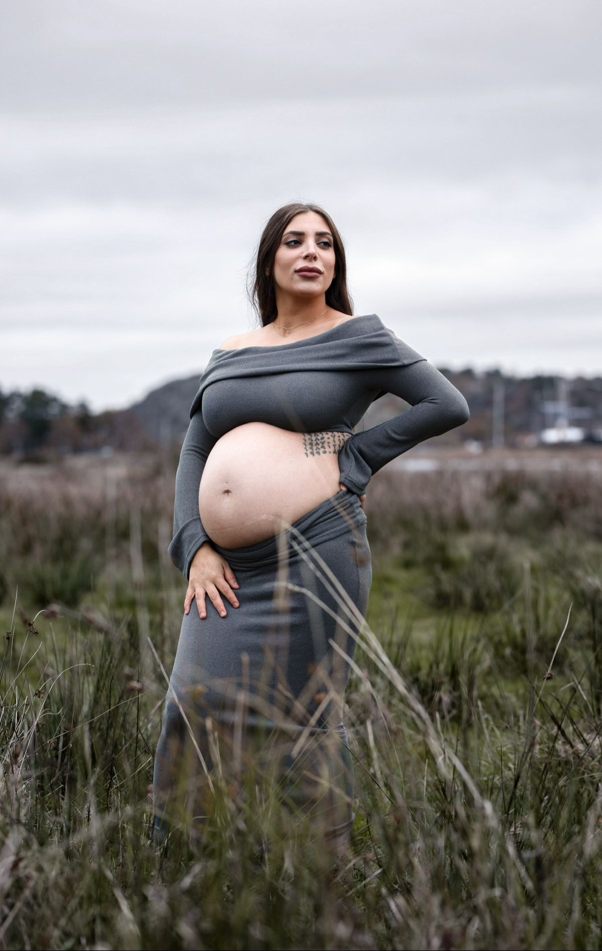 gravid kvinna står på en åker. på sig har hon ett grått stickat set bestående av en långärmad topp och en kjol.