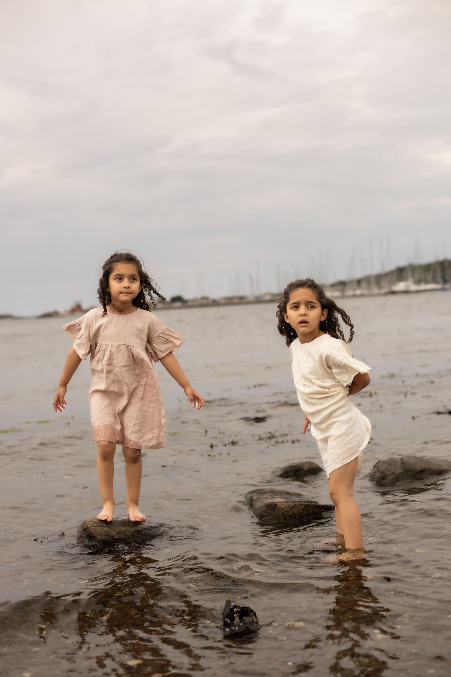 två barn står i havet, ena står på en sten. de har båda klänningar på sig. en rosa och en vit.