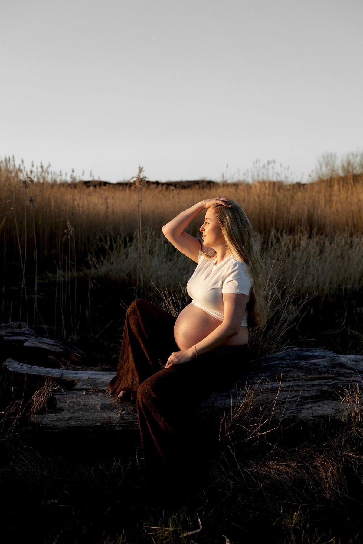 Gravid kvinna sitter på en stock. på sig har hon en vit topp och ett par bruna byxor.