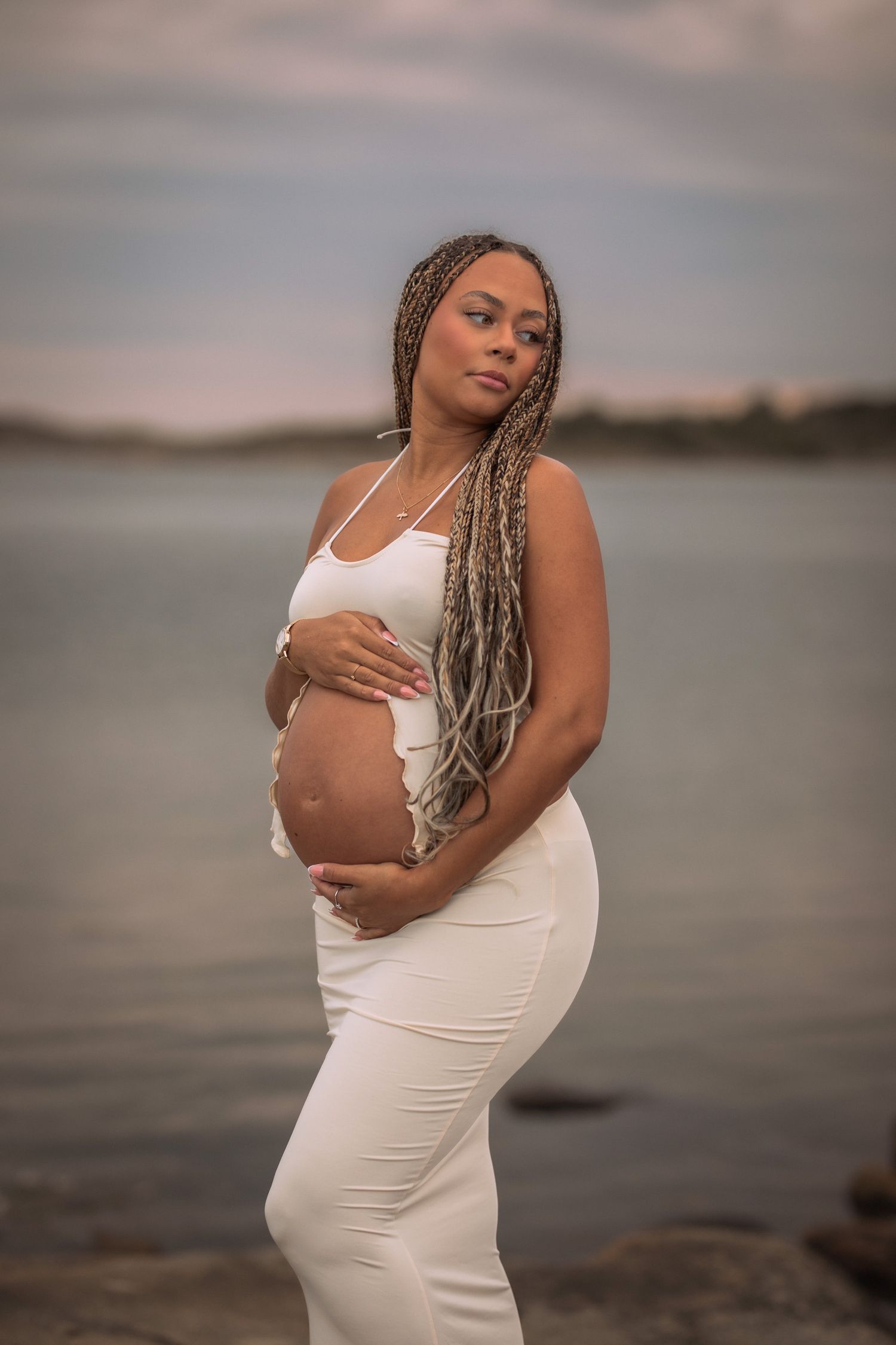 gravid kvinna står med havet bakom sig. hon håller om sin mage. på sig har hon ett vitt set bestående av en kjol och topp.