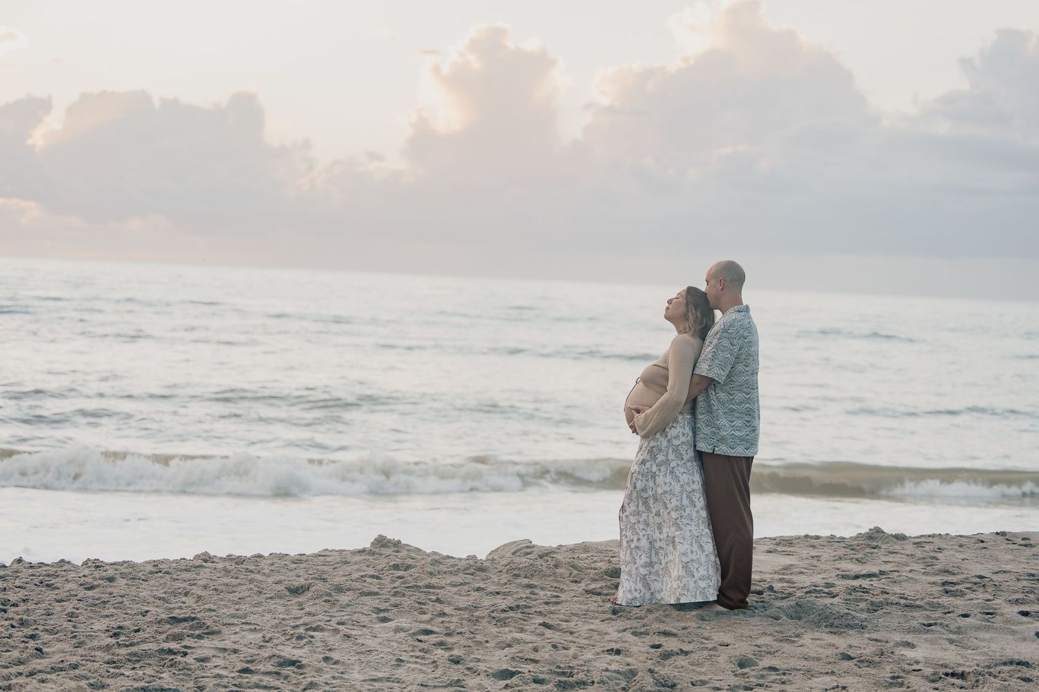 Couple embracing on sandy beach at sunset with gentle ocean waves and pastel cloudy sky in background.