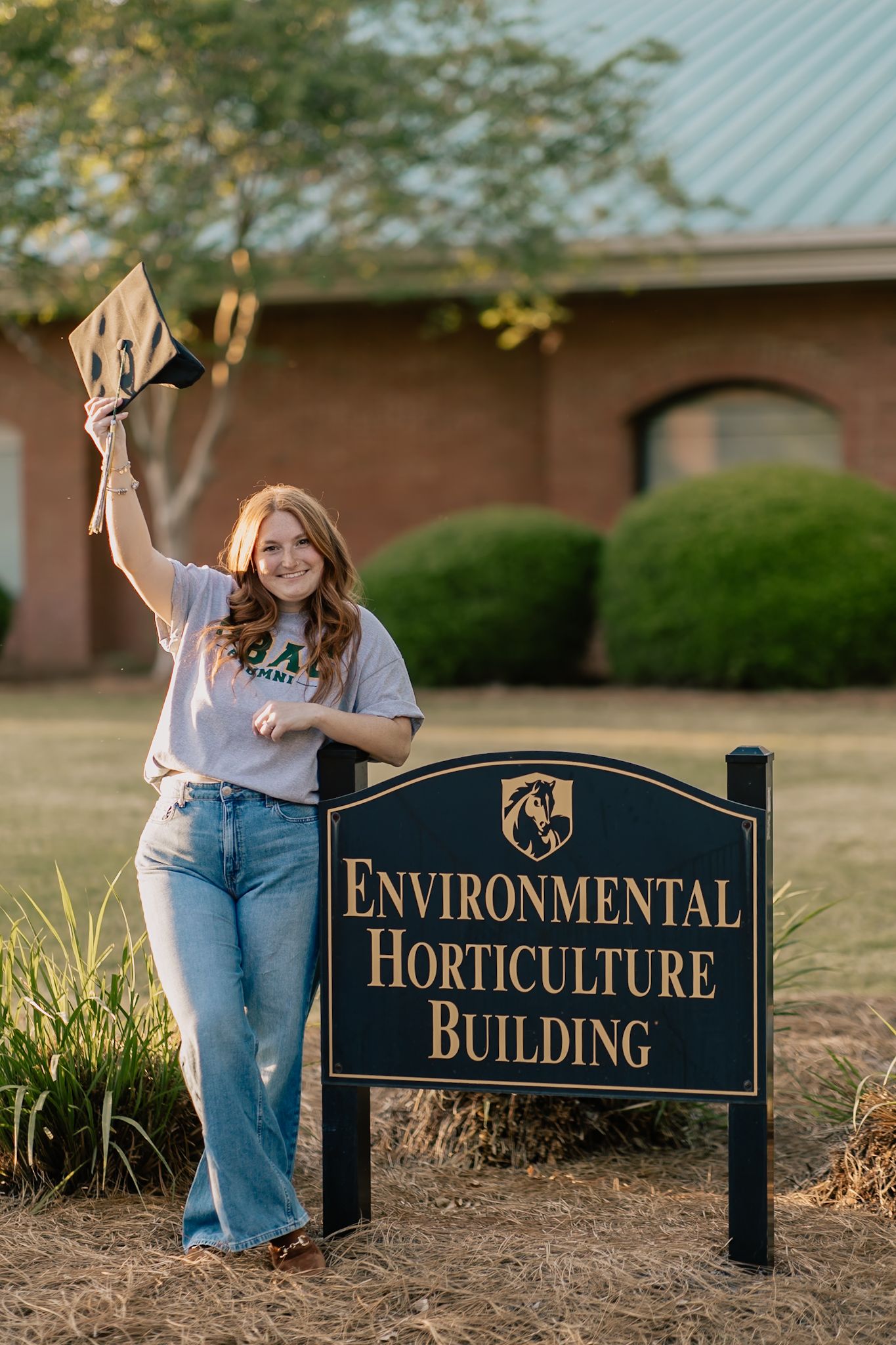 A smiling young woman holding a diploma beside an Environmental Horticulture Building sign outdoors.