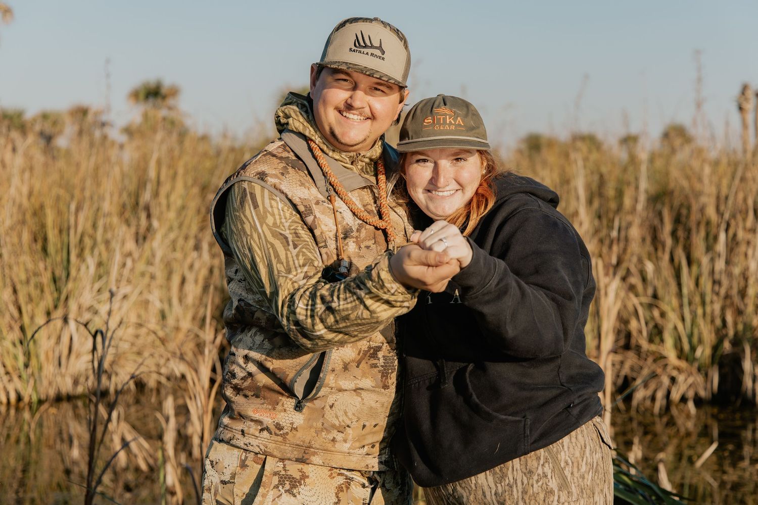A smiling couple in camouflage gear embracing outdoors near a marsh during a hunting trip.