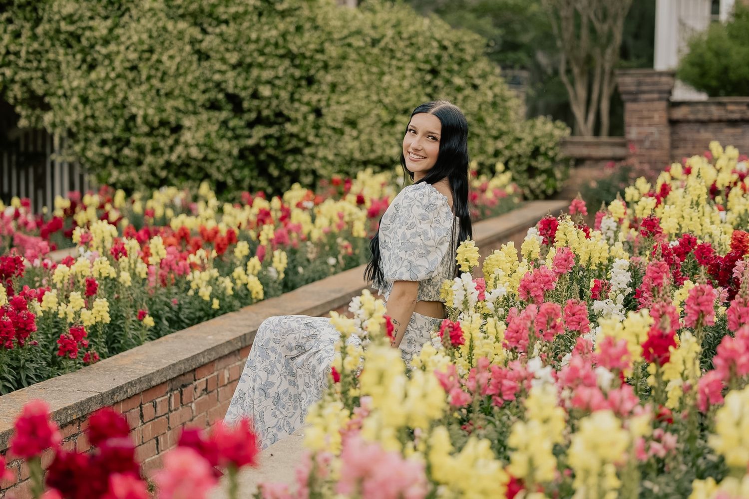 A woman in a floral dress sitting among vibrant pink, red, and yellow snapdragon flowers in a garden.