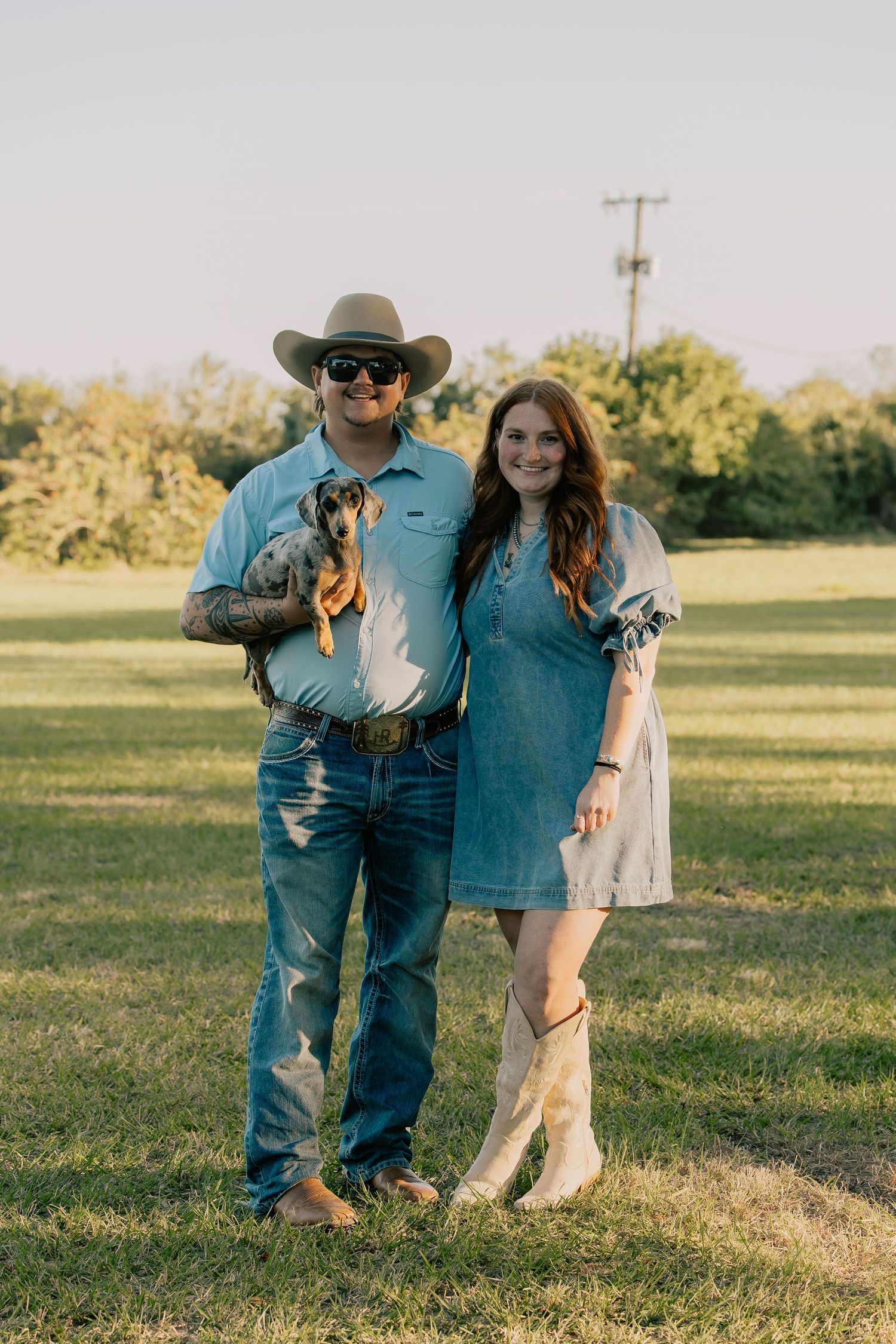 A couple in western attire holding a dachshund puppy while standing in a sunny green field.