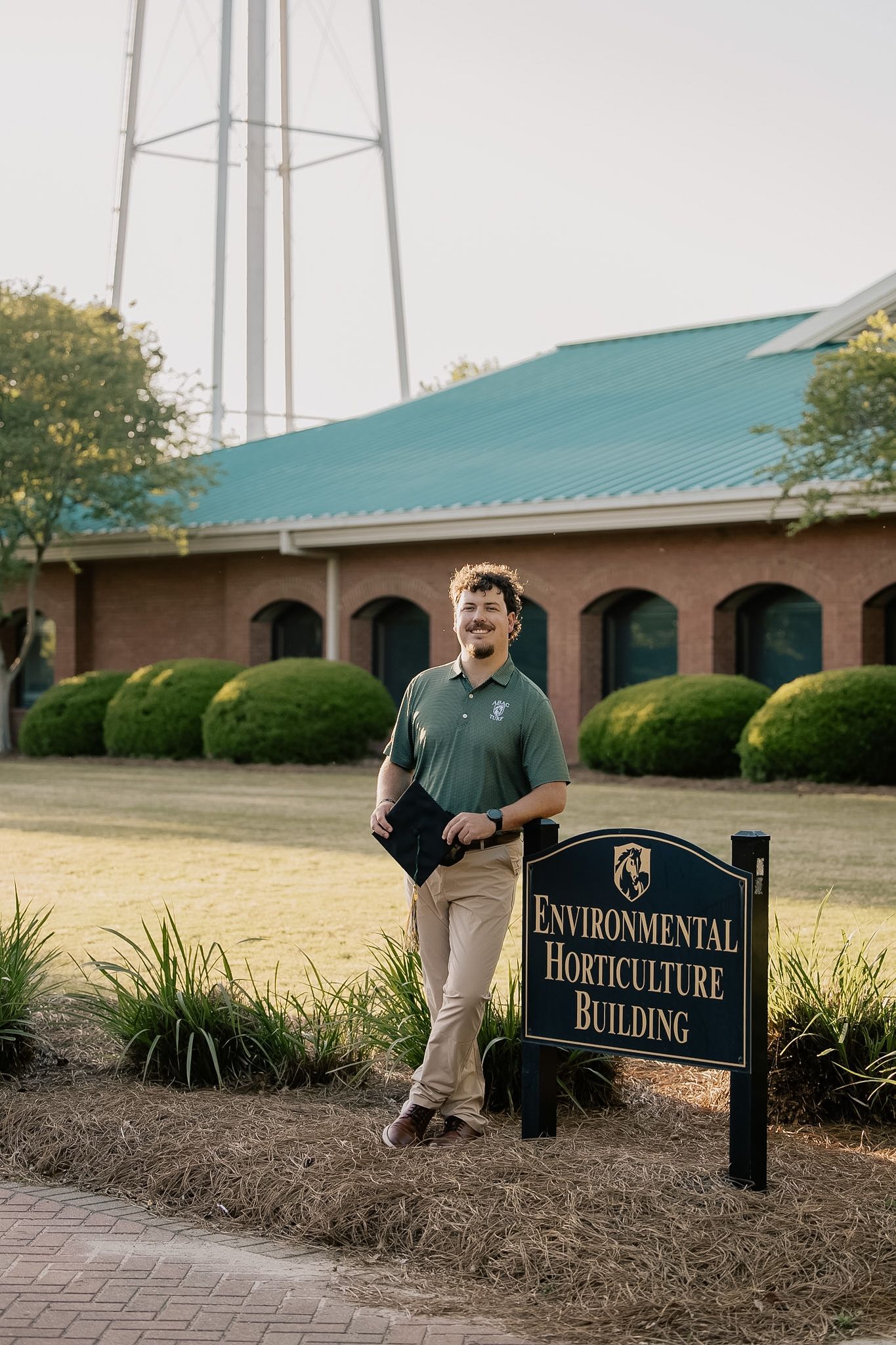 A cheerful young man laughs while standing next to the Environmental Horticulture Building sign on campus.