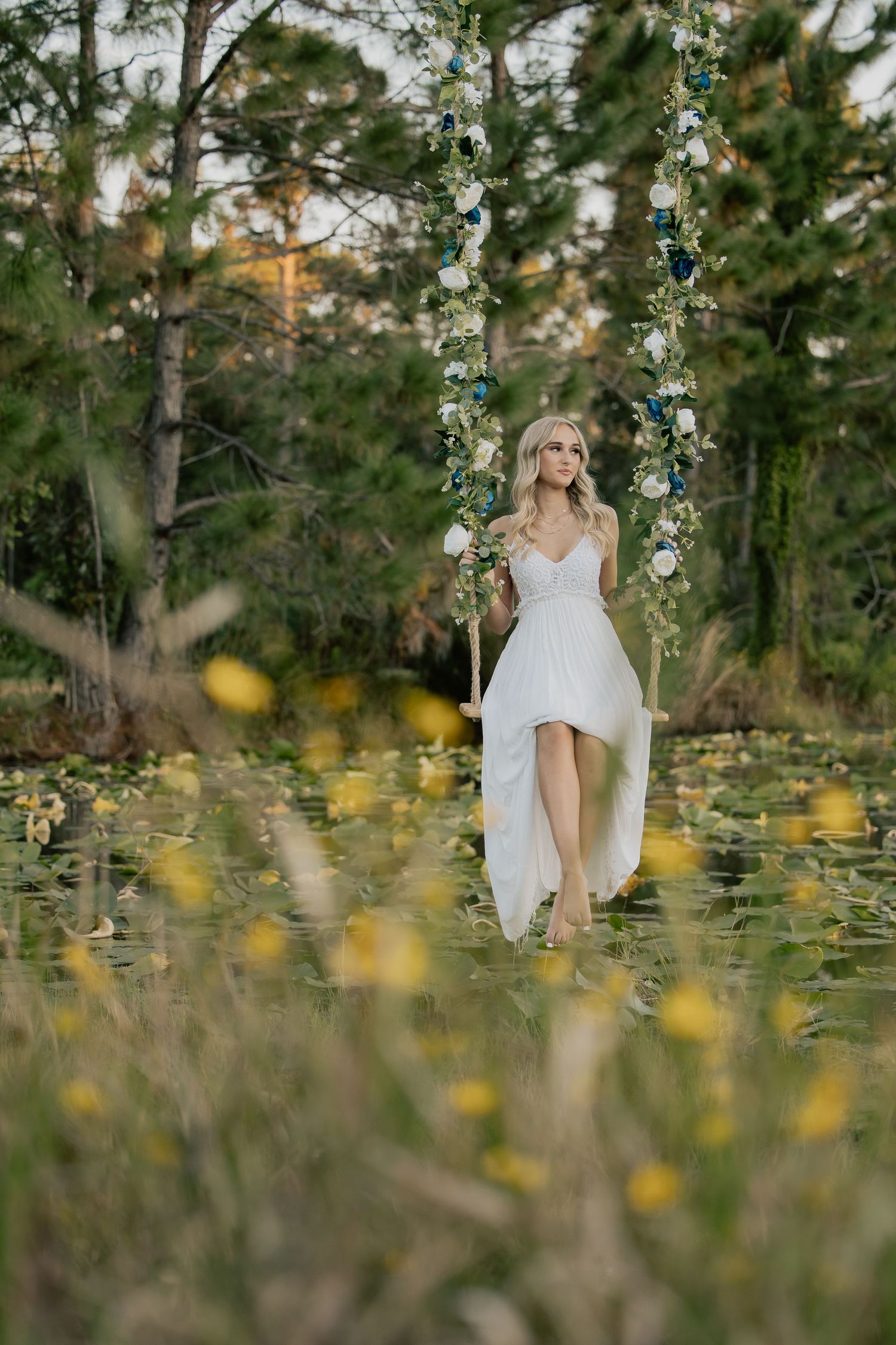 Blonde bride in white gown sitting on floral swing surrounded by yellow wildflowers at sunset.