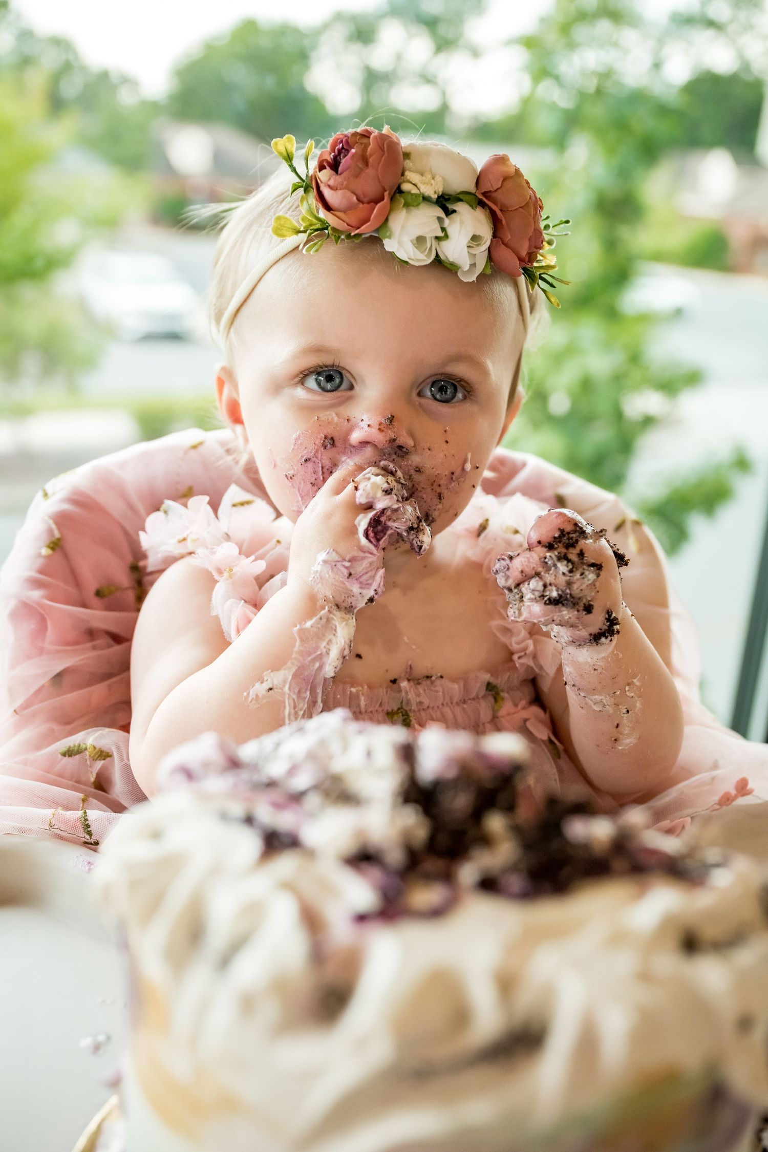 Baby girl in floral crown eating first birthday cake, face covered in frosting, sitting in high chair.