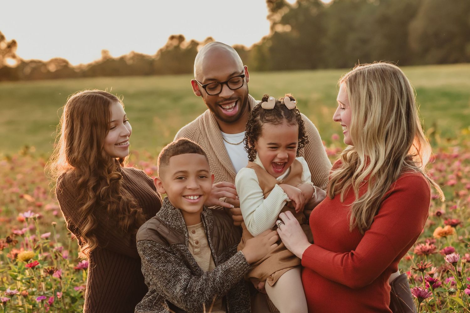 Happy family of five laughing together in a sunlit flower field during golden hour outdoor portrait session.