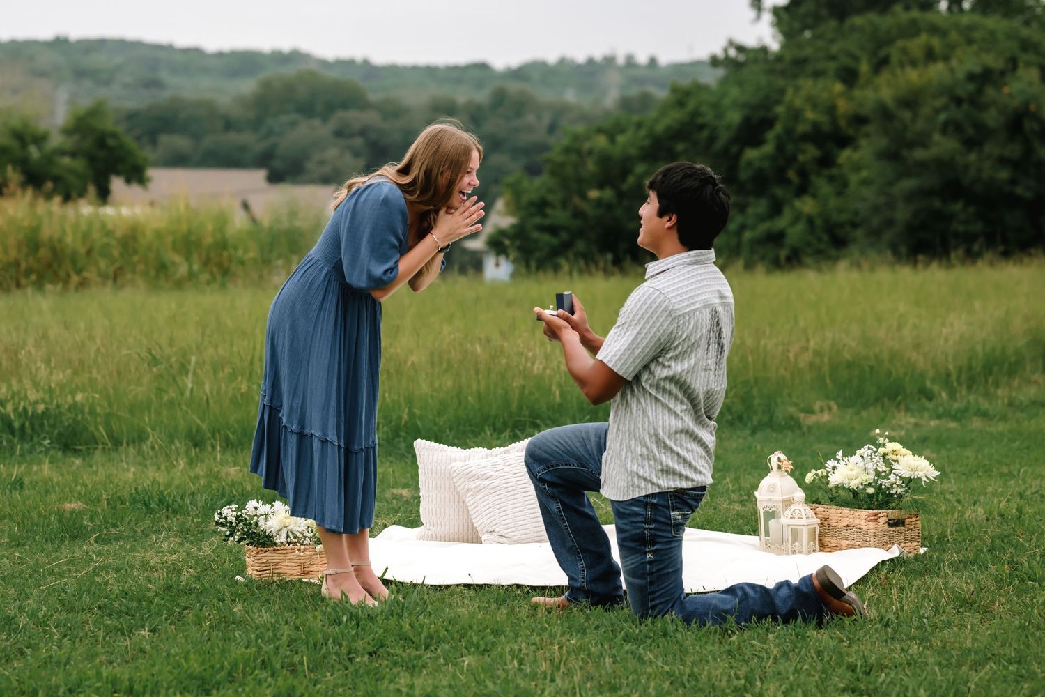 Two people enjoy a romantic picnic on a lush green hillside with a blanket and basket spread out on the grass.