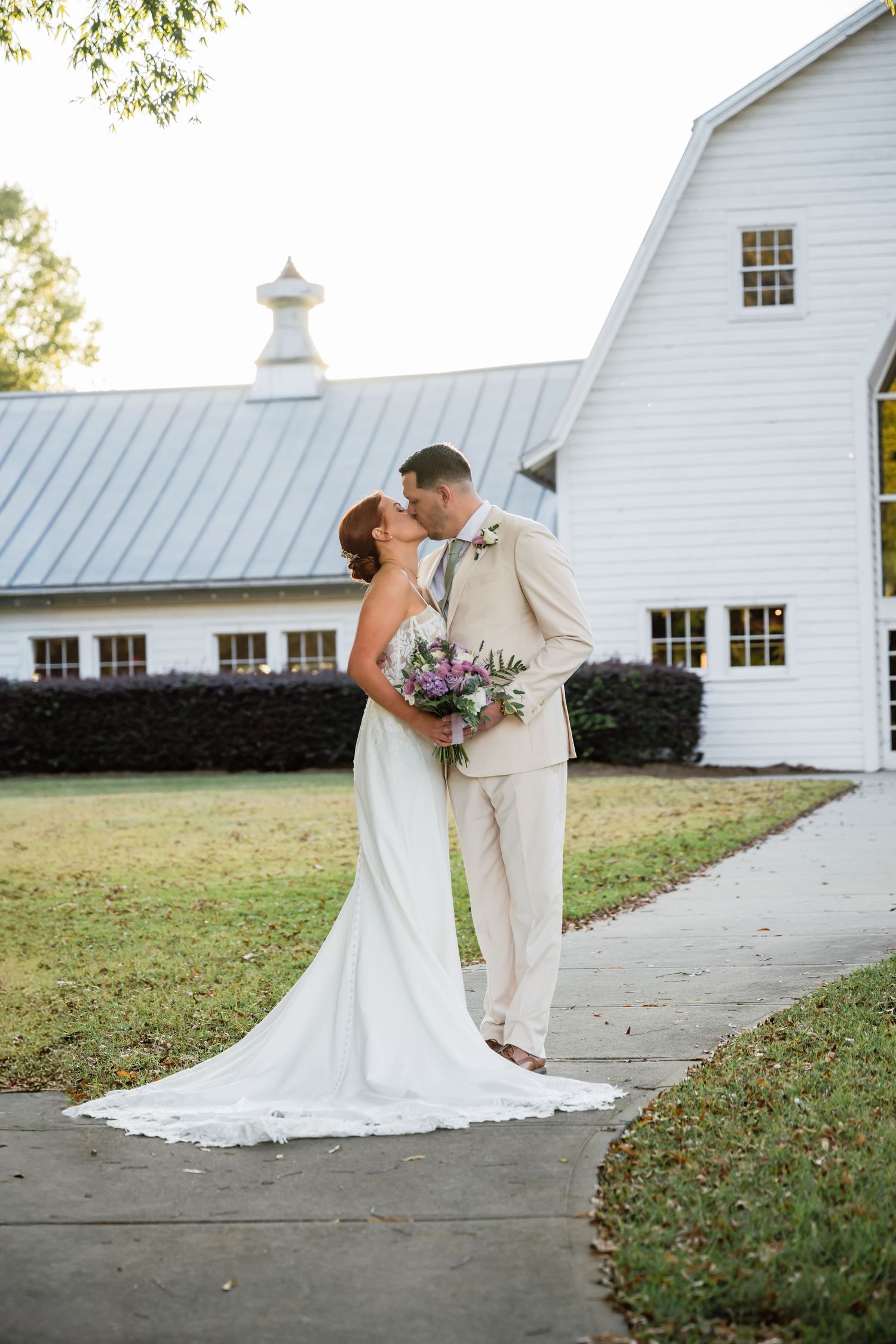 A romantic wedding couple shares a kiss in front of a white barn building on a grassy lawn.