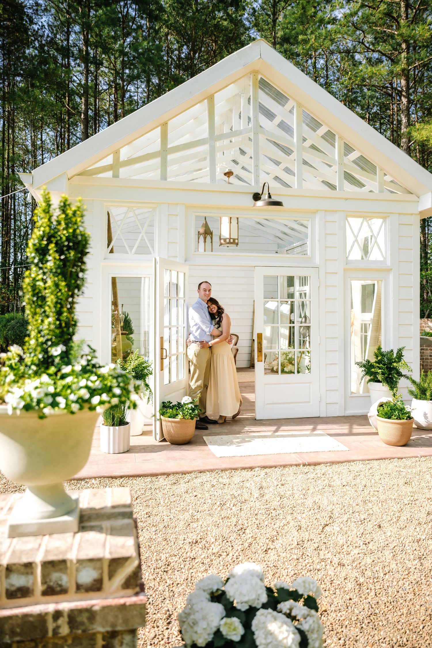 A white garden greenhouse structure with French doors and potted plants in a wooded setting.