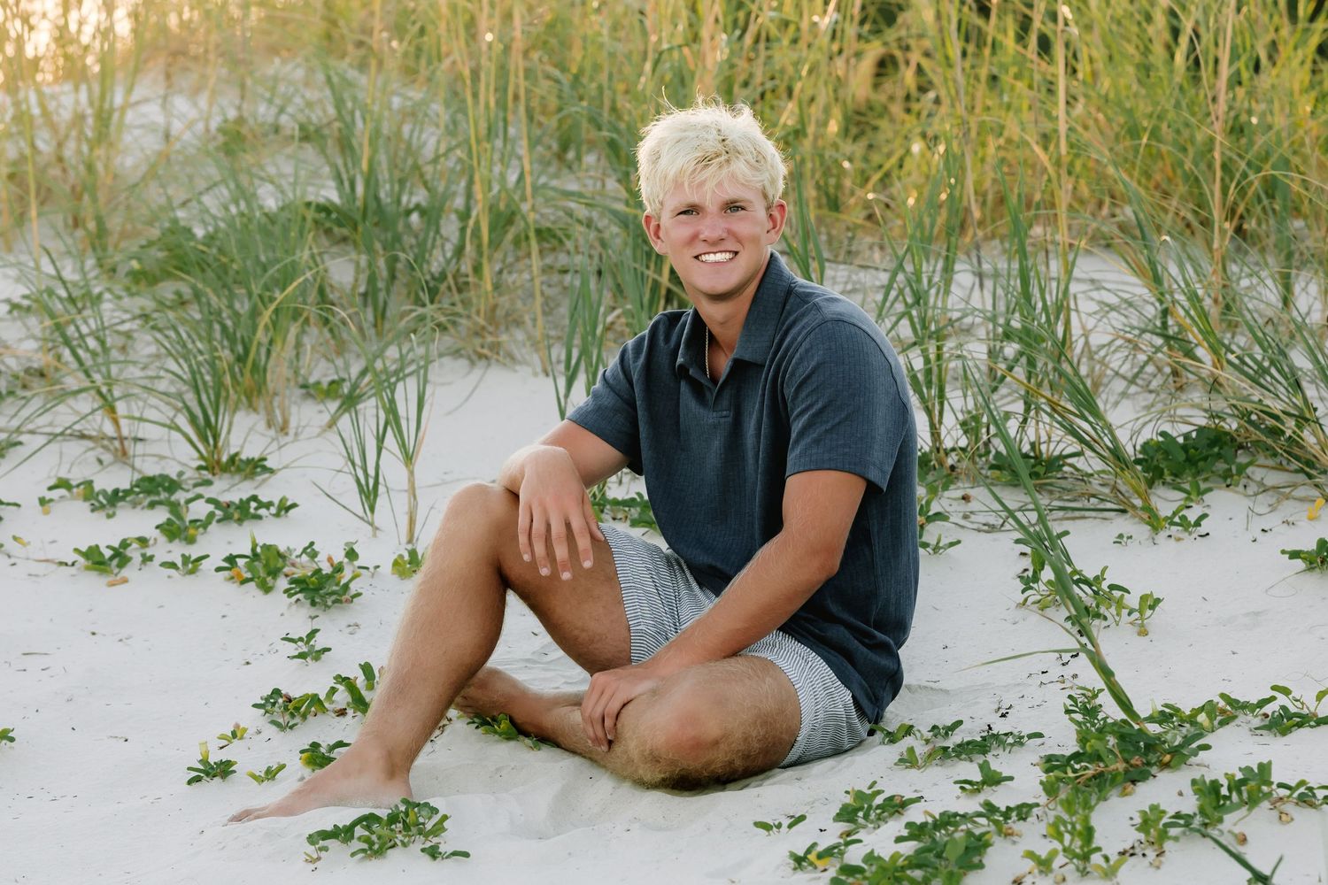 Person sits casually on white sand beach with sea oats and coastal vegetation at sunset.