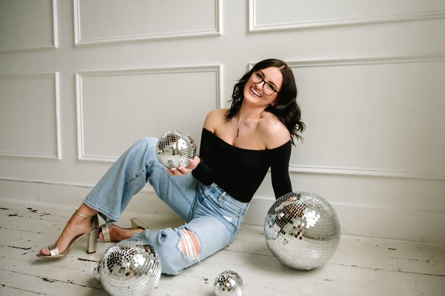 A woman in a black top and jeans poses on a white floor with silver disco balls around her.