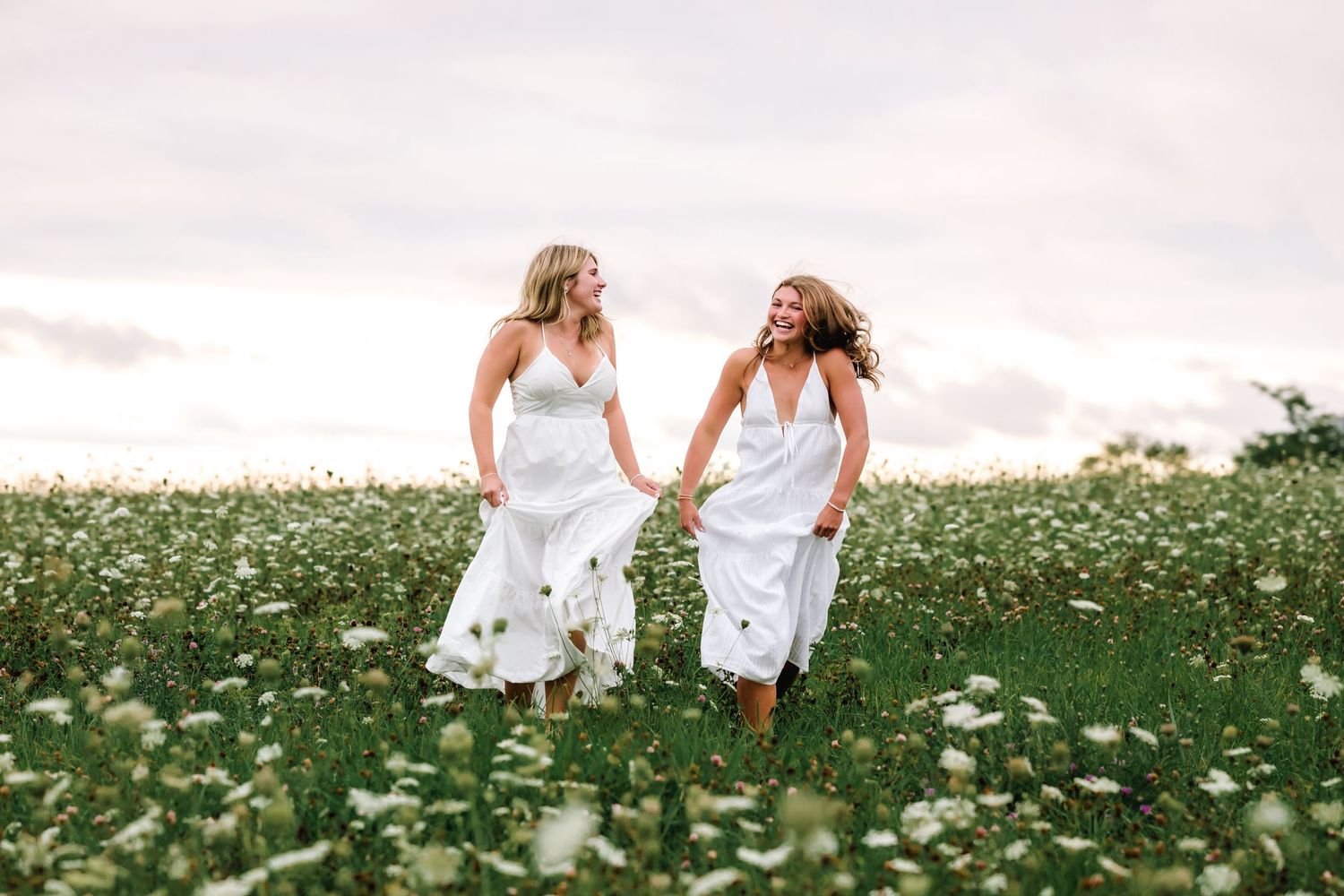 Two people in white dresses run playfully through a blooming wildflower meadow on a cloudy summer day.