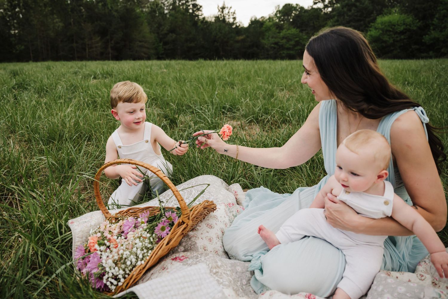 Mother sitting on blanket in field, holding baby while offering flower to toddler with picnic basket.