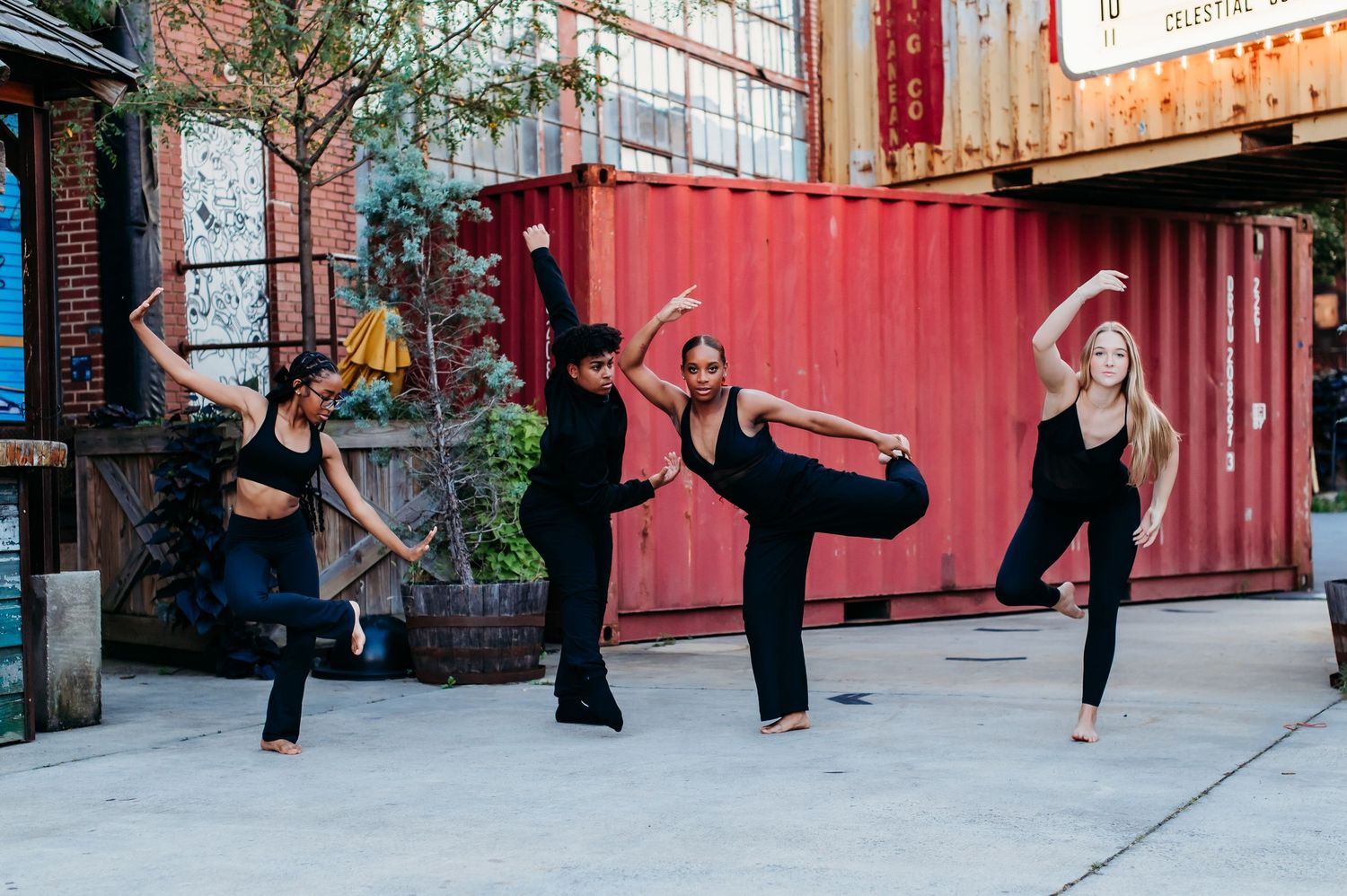 Four dancers in black attire strike synchronized poses against an urban backdrop with red shipping container.