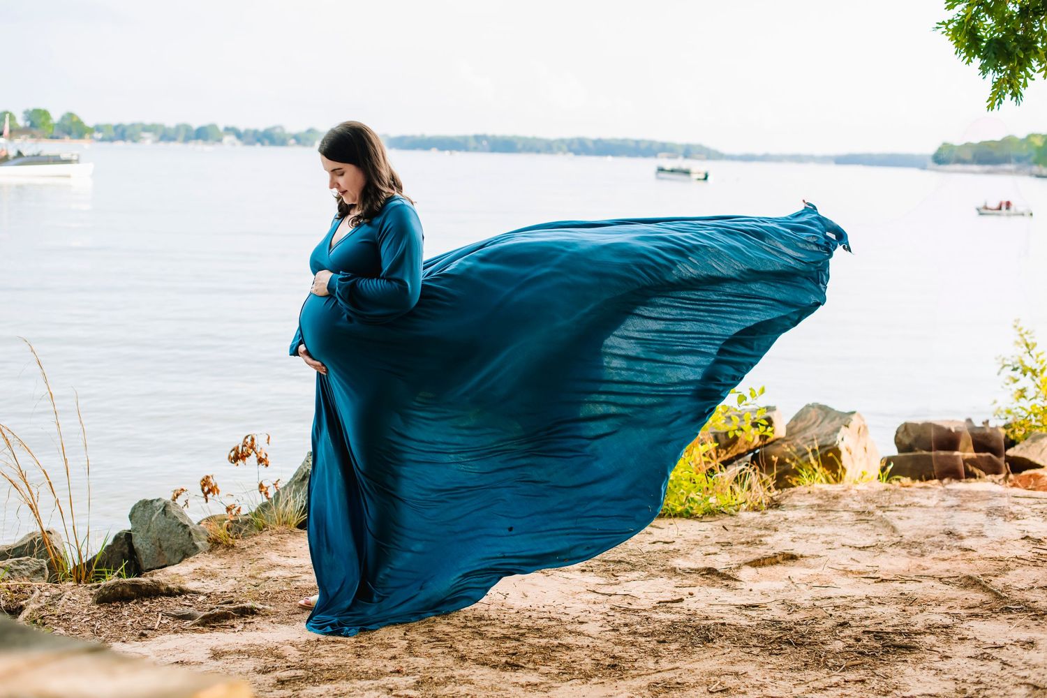 A pregnant woman in a flowing teal dress stands by a lake with her gown billowing in the wind.