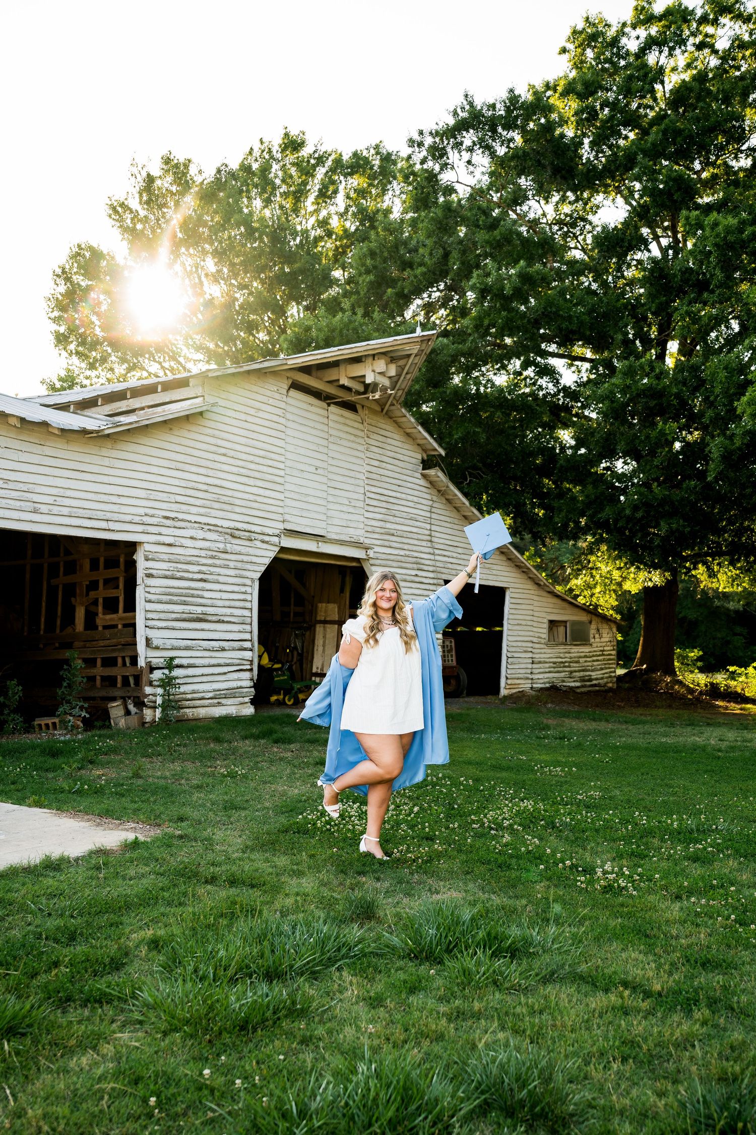 Graduate in blue cap and gown celebrates outdoors near rustic barn at golden hour.