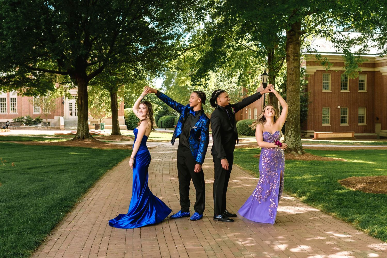 A wedding party poses for photos on a tree-lined pathway at a historic brick building campus.