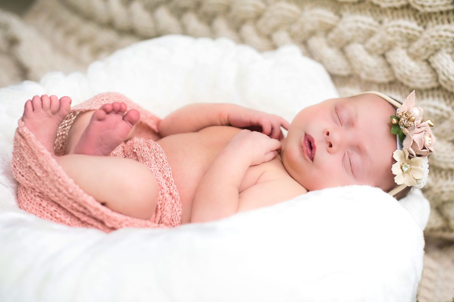 A sleeping newborn baby wearing a pink wrap lies on white bedding with floral accessories.