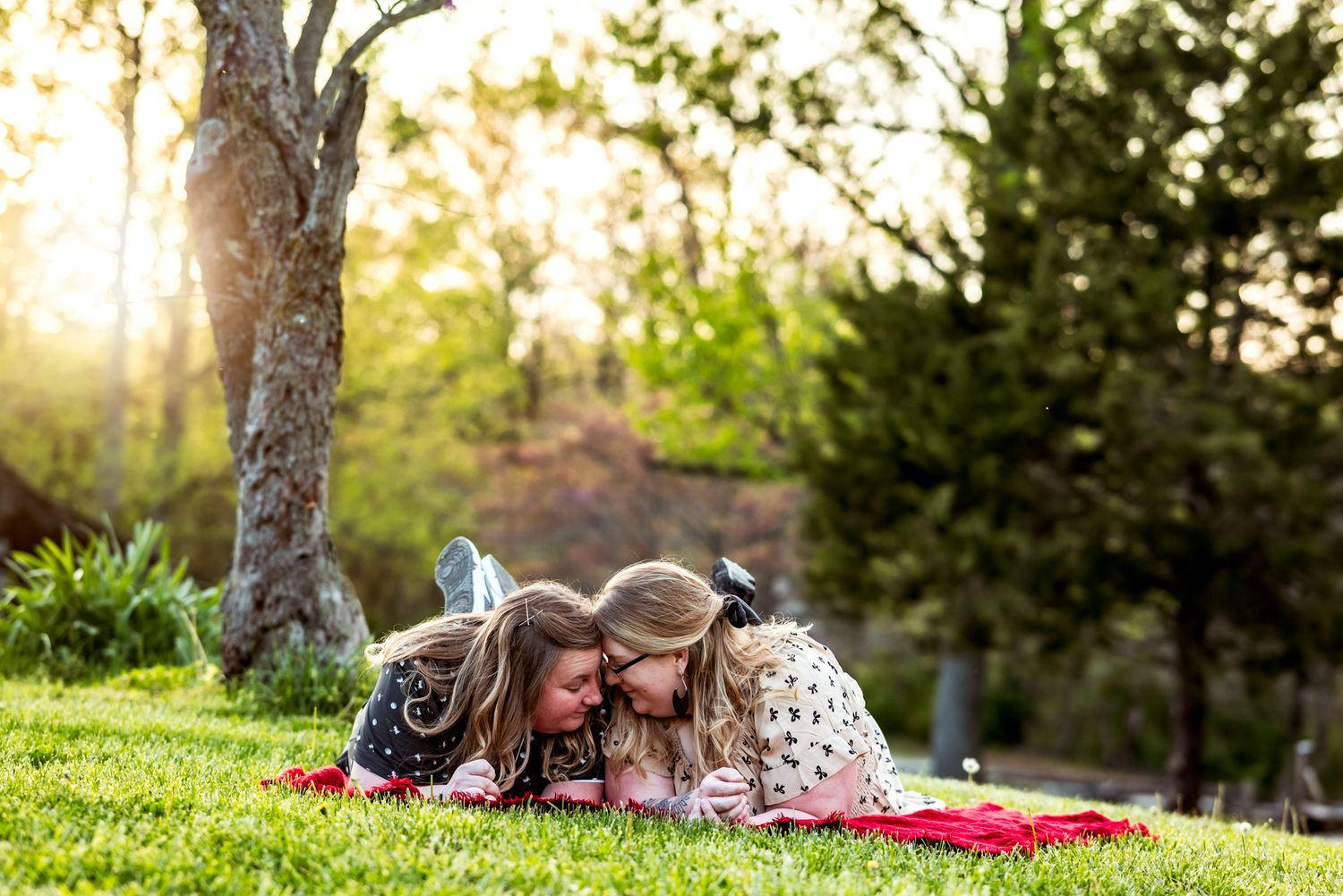 Two children laying on a red blanket reading together on a grassy hill at sunset in a park.