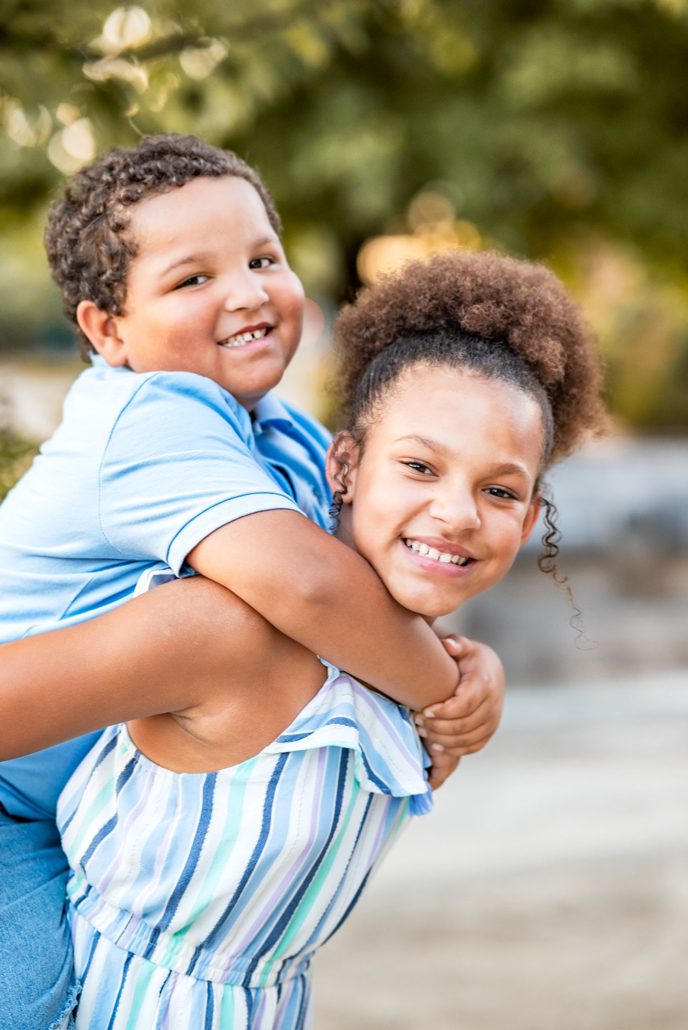 Two young siblings sharing a playful piggyback ride while wearing light blue summer outfits.