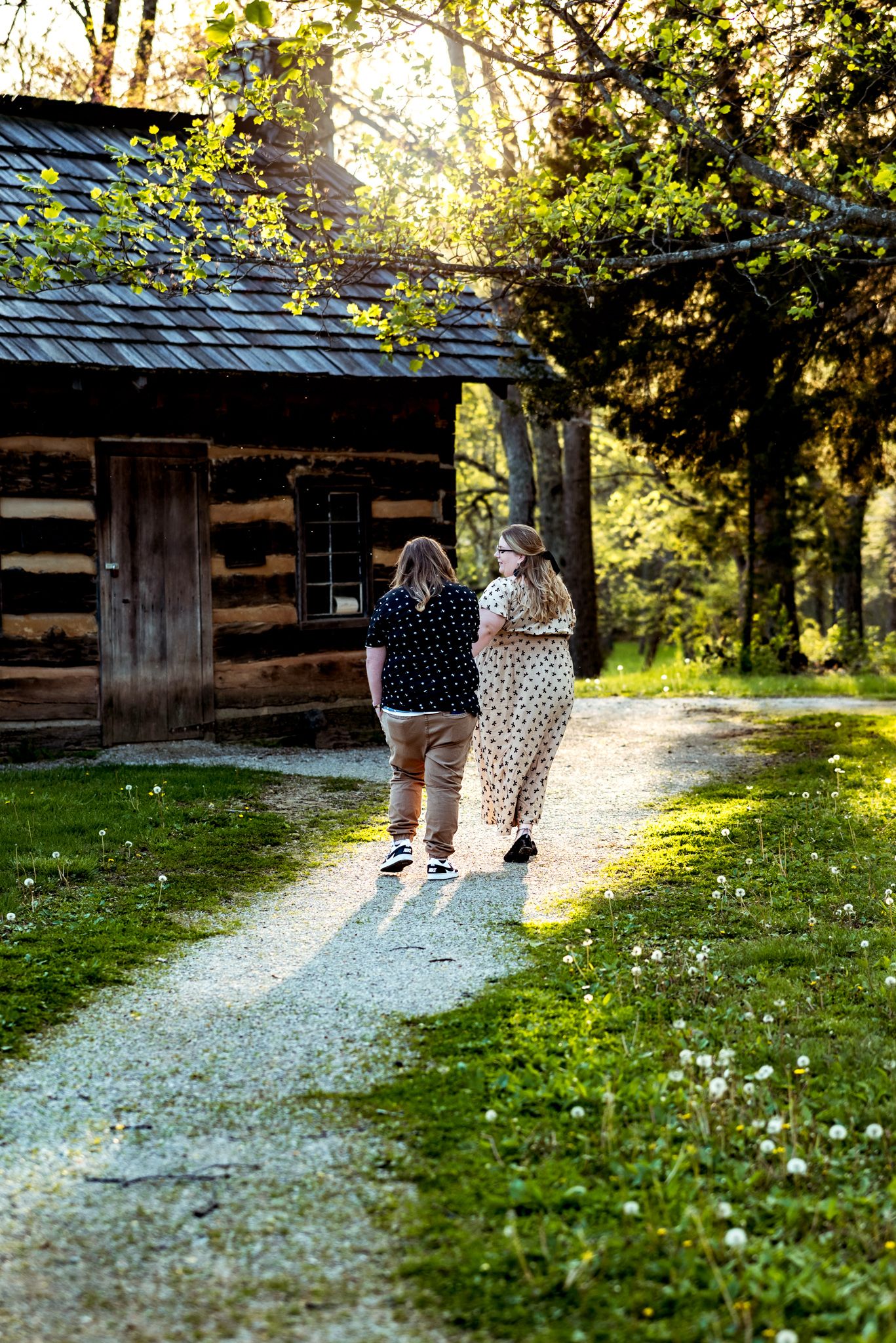 Two people walk together on a gravel path near a rustic log cabin as sunlight filters through trees.
