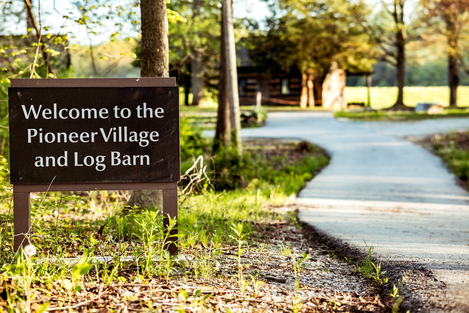 Welcome sign for Pioneer Village and Log Barn along a winding pathway surrounded by trees and sunlit grass.