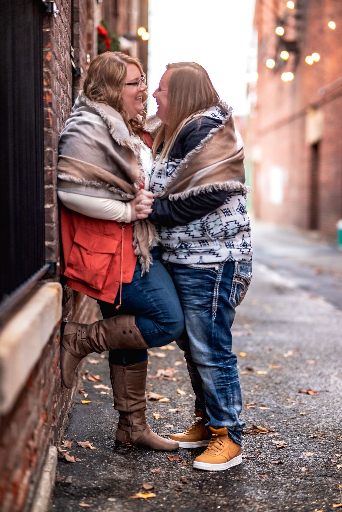 Two people share an intimate moment laughing together while standing against a brick wall on an autumn day.