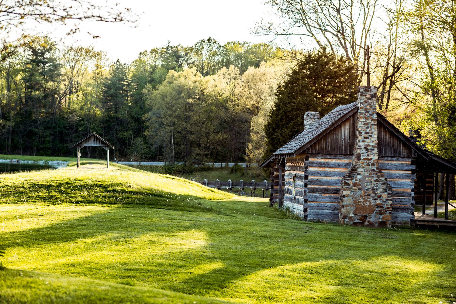 Rustic log cabin sits on a sunlit grassy meadow with a wooden gazebo in the background during golden hour.