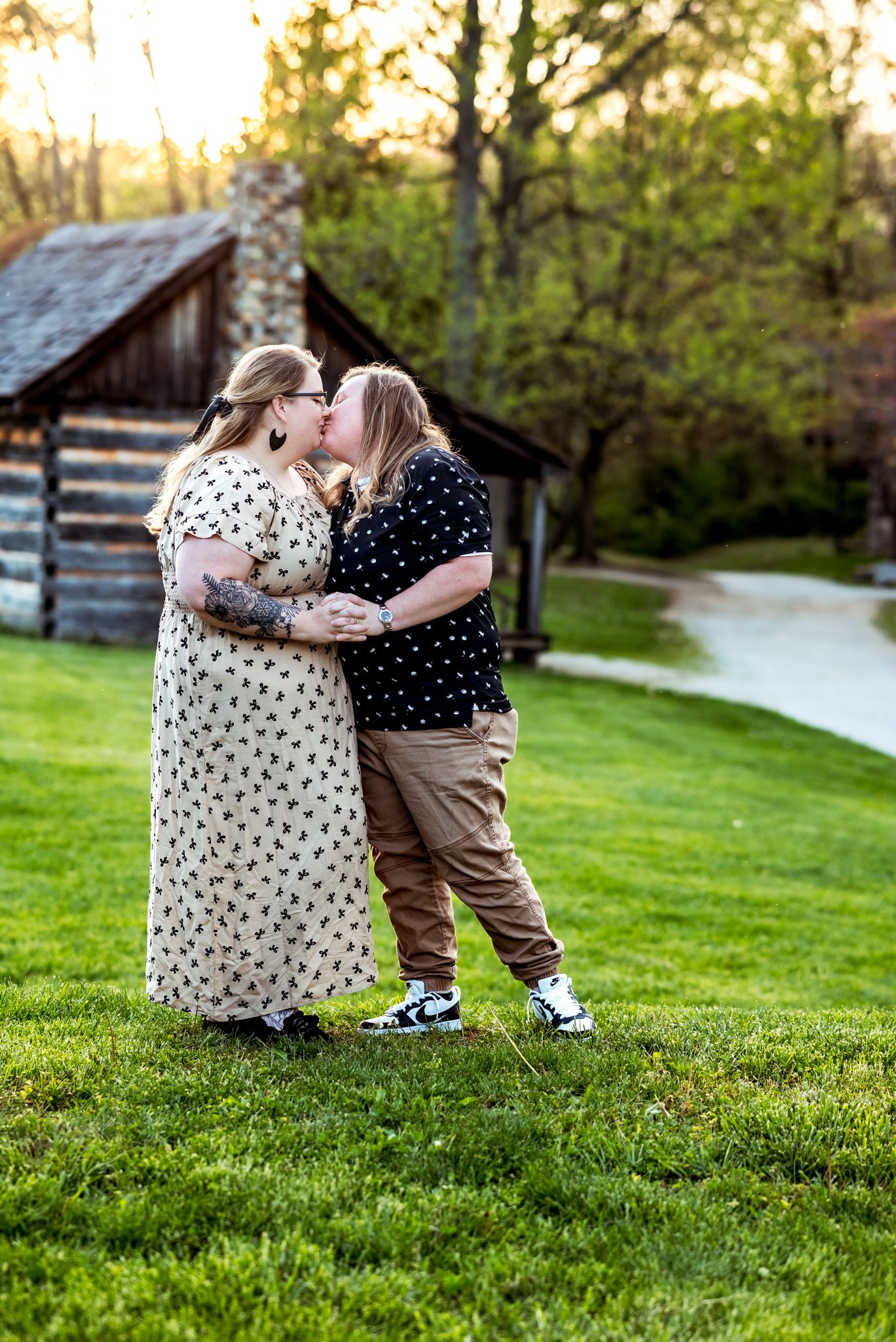 A couple in matching polka dot dresses embrace on a grassy lawn with a log cabin in the background.