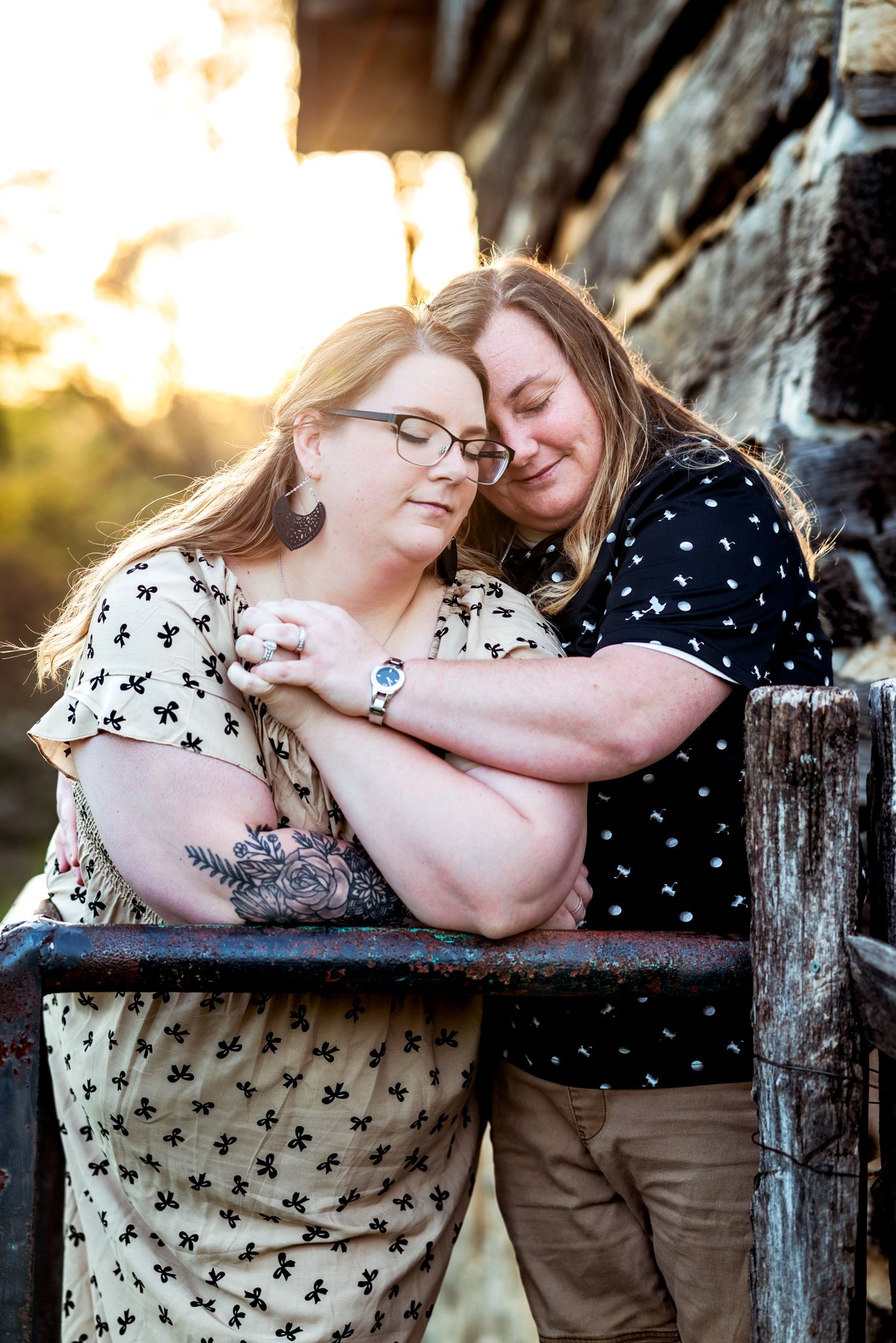 Two people share an intimate embrace while leaning on a rustic wooden railing at sunset.